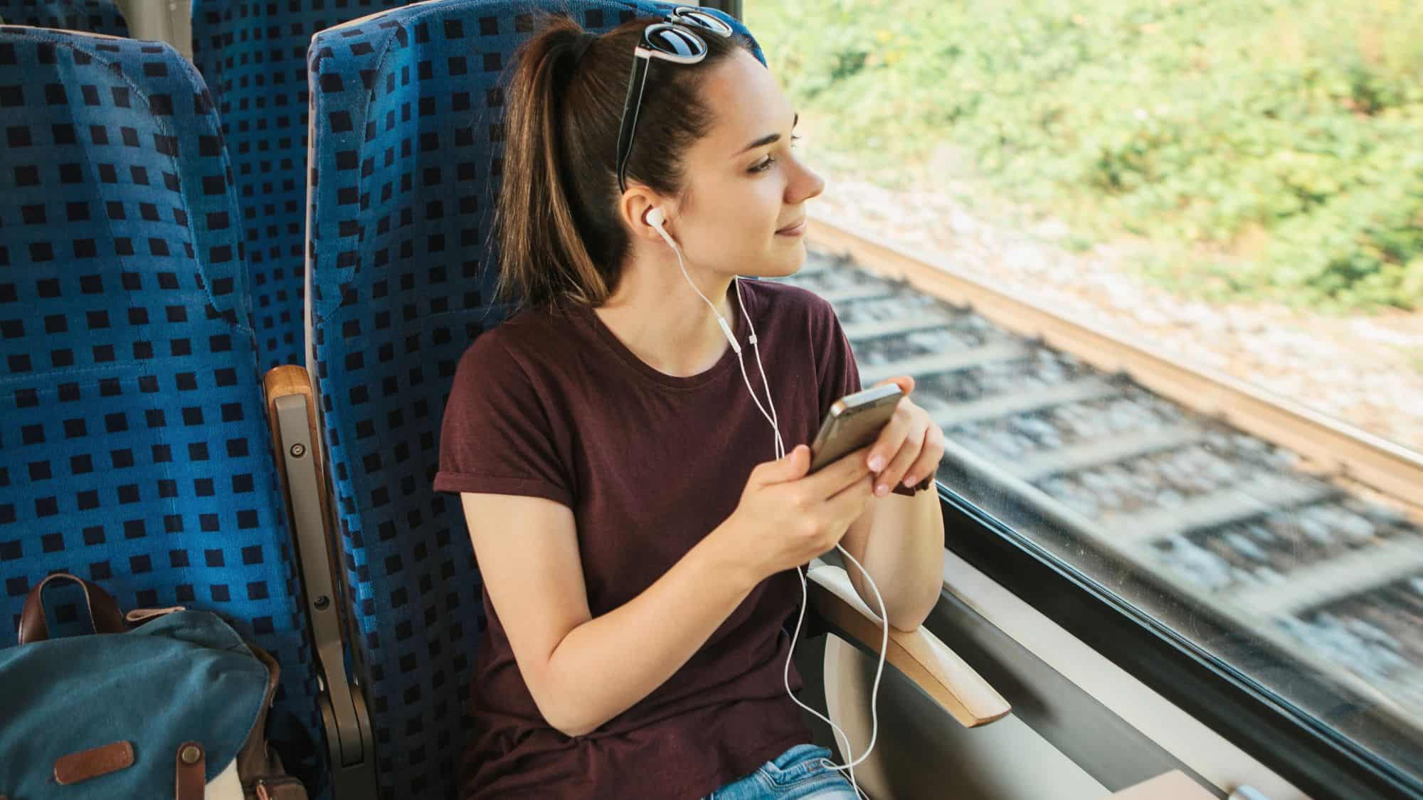 A young woman with sunglasses resting on her head listens to music with earbuds while looking out the window of a train. She holds a smartphone in her hand, enjoying the journey with the train tracks visible outside.