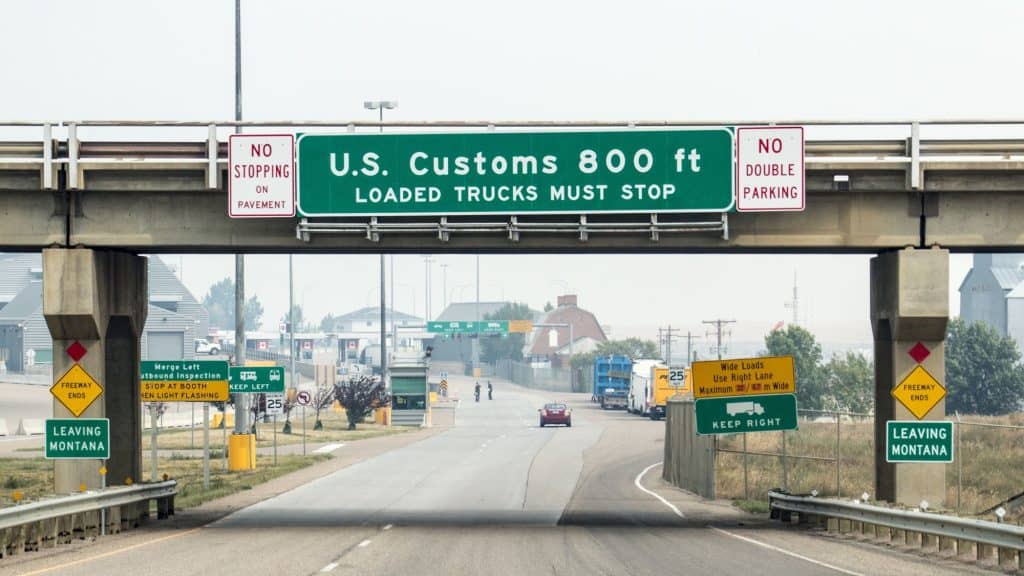 A highway leading to a U.S. border checkpoint, marked by a large overhead sign reading "U.S. Customs 800 ft - Loaded Trucks Must Stop." Various road signs indicate direction and traffic rules, with "Leaving Montana" signs visible.