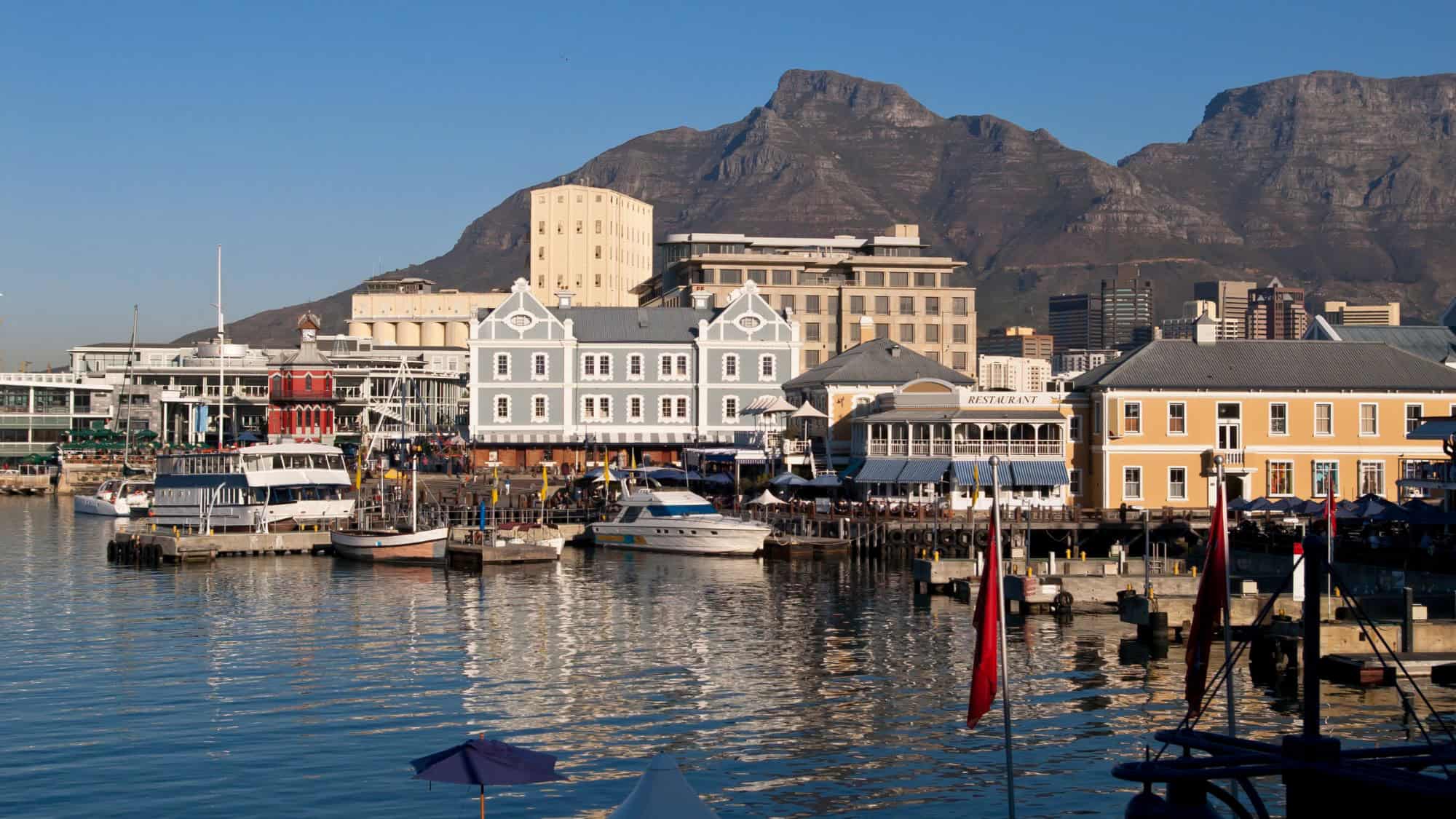 A vibrant harbor scene with colorful historic buildings, boats, and a lively waterfront atmosphere. Table Mountain looms in the background, providing a stunning urban-meets-nature backdrop.