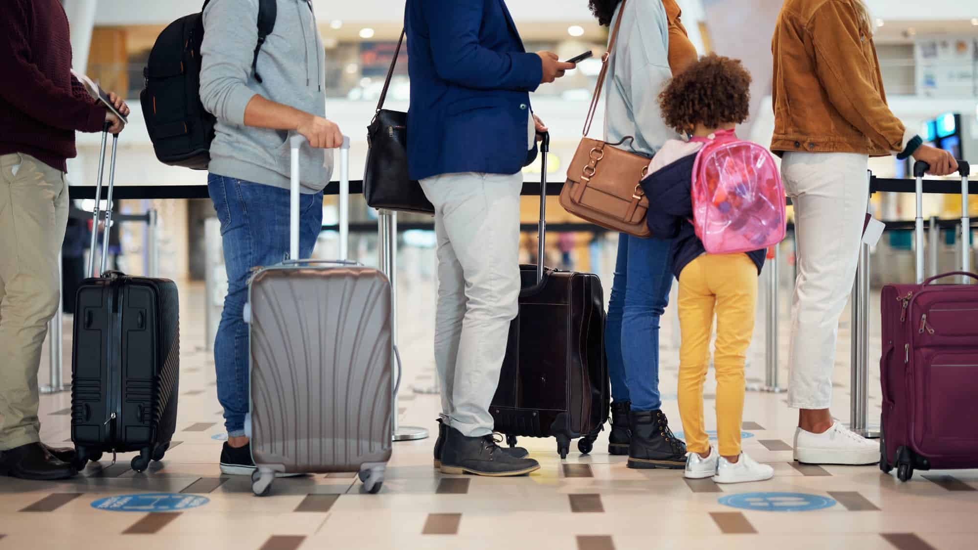A group of travelers stands in line at an airport, pulling rolling suitcases. A young girl with a pink backpack and yellow pants stands among them, looking ahead while waiting her turn.