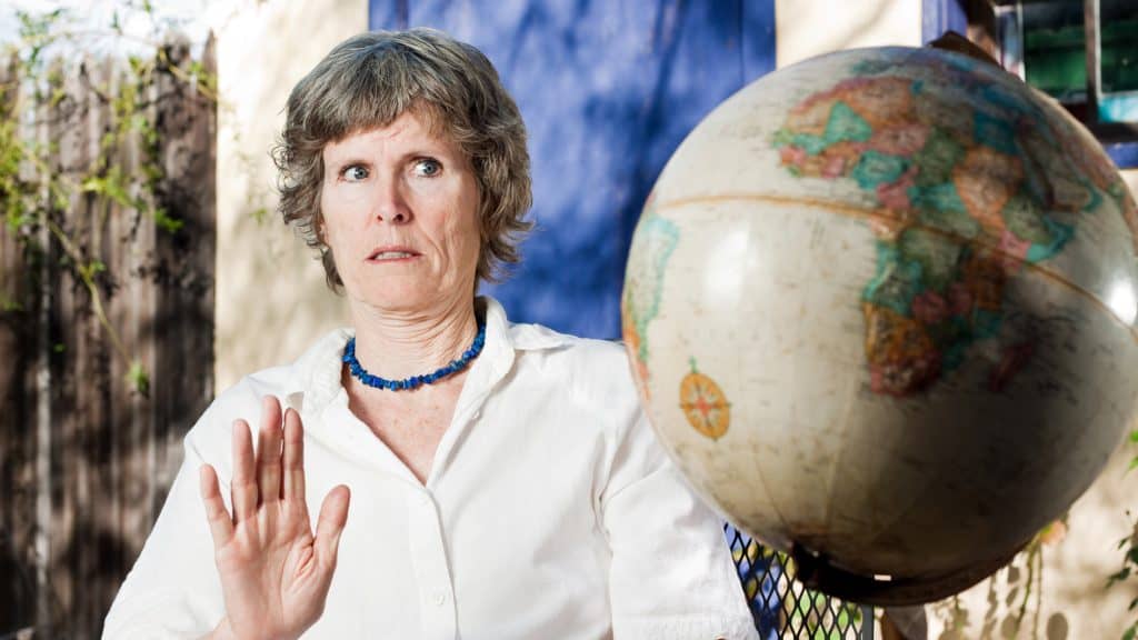 A skeptical-looking older woman in a white blouse and blue beaded necklace gestures "no" while sitting outdoors, with a large spinning globe in front of her.
