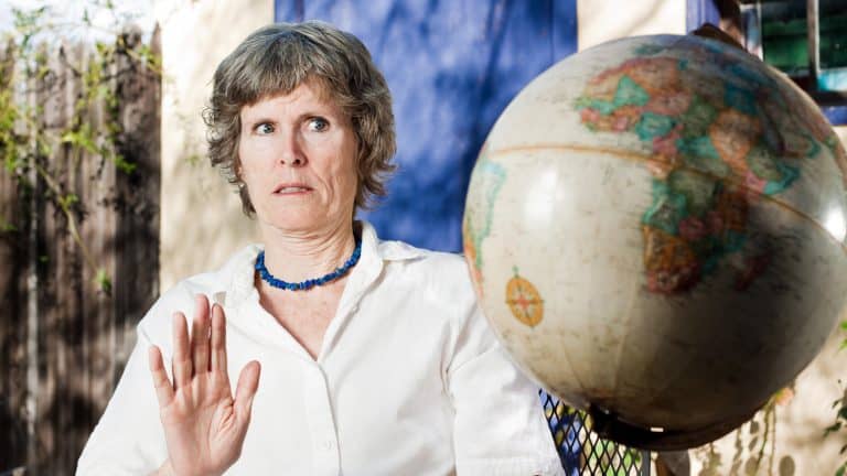 A skeptical-looking older woman in a white blouse and blue beaded necklace gestures "no" while sitting outdoors, with a large spinning globe in front of her.