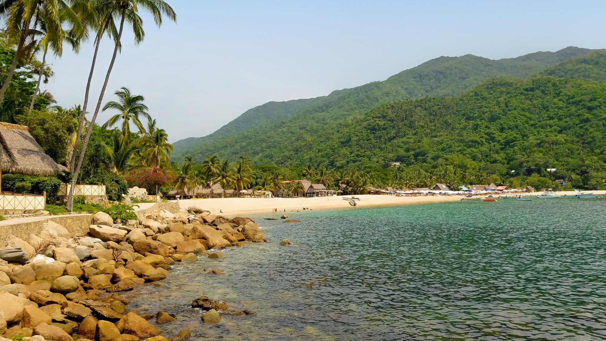 A serene beach in Yelapa, Mexico, with golden sand, rocky shores, and lush green hills in the background, framed by tall palm trees and quaint beachfront huts.