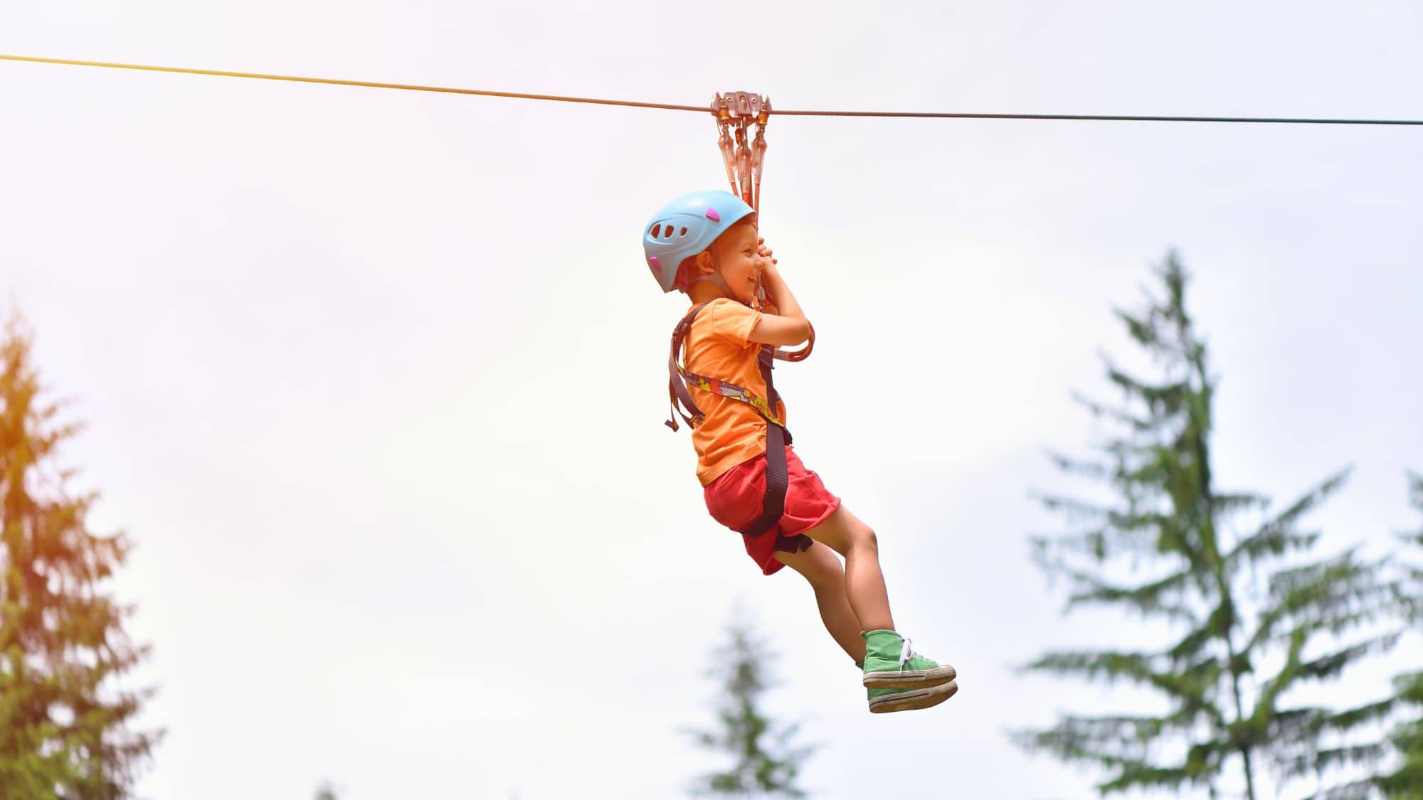 A young child wearing a blue helmet and safety harness is mid-air on a zip line, gripping the harness and smiling. The background is slightly blurred with tall evergreen trees visible.