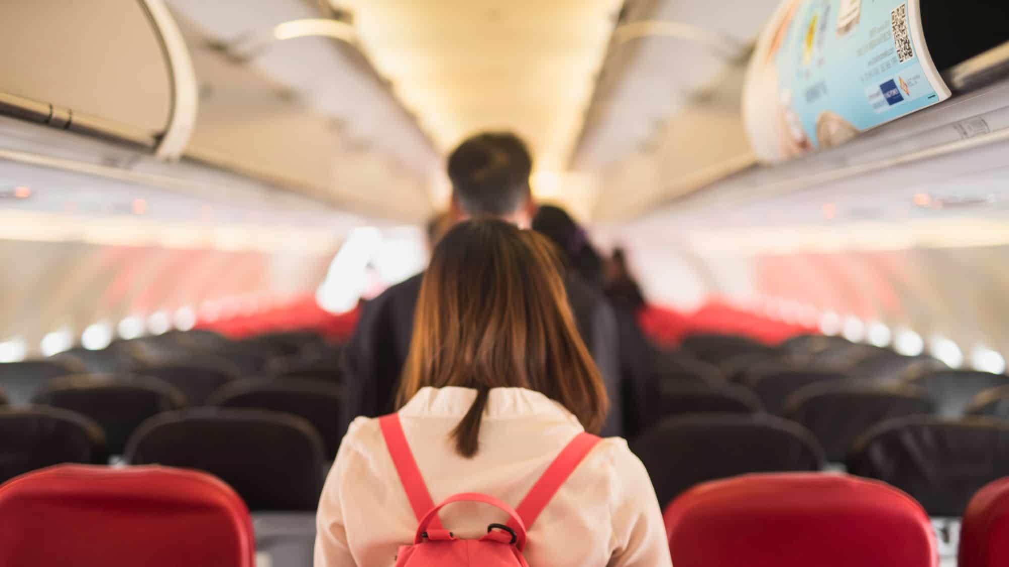 A traveler with a pink backpack stands in a crowded airplane aisle, waiting to exit while other passengers gather ahead. The red and black seats are empty, creating a tunnel-like view toward the front of the plane.