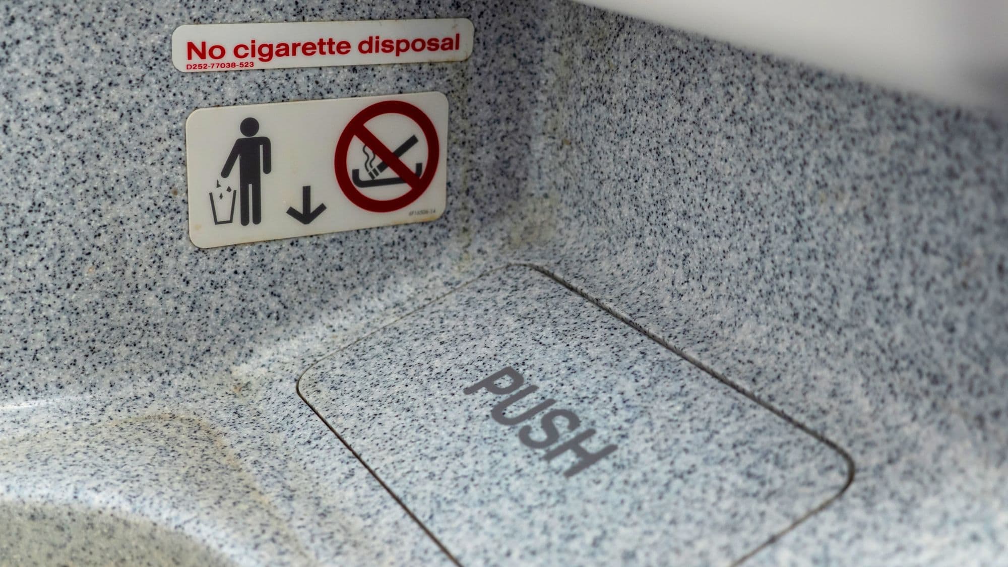 Close-up of a speckled gray airplane lavatory trash bin with a sign warning "No cigarette disposal" and a push flap for trash. The signage includes icons for proper waste disposal and a red crossed-out cigarette symbol.