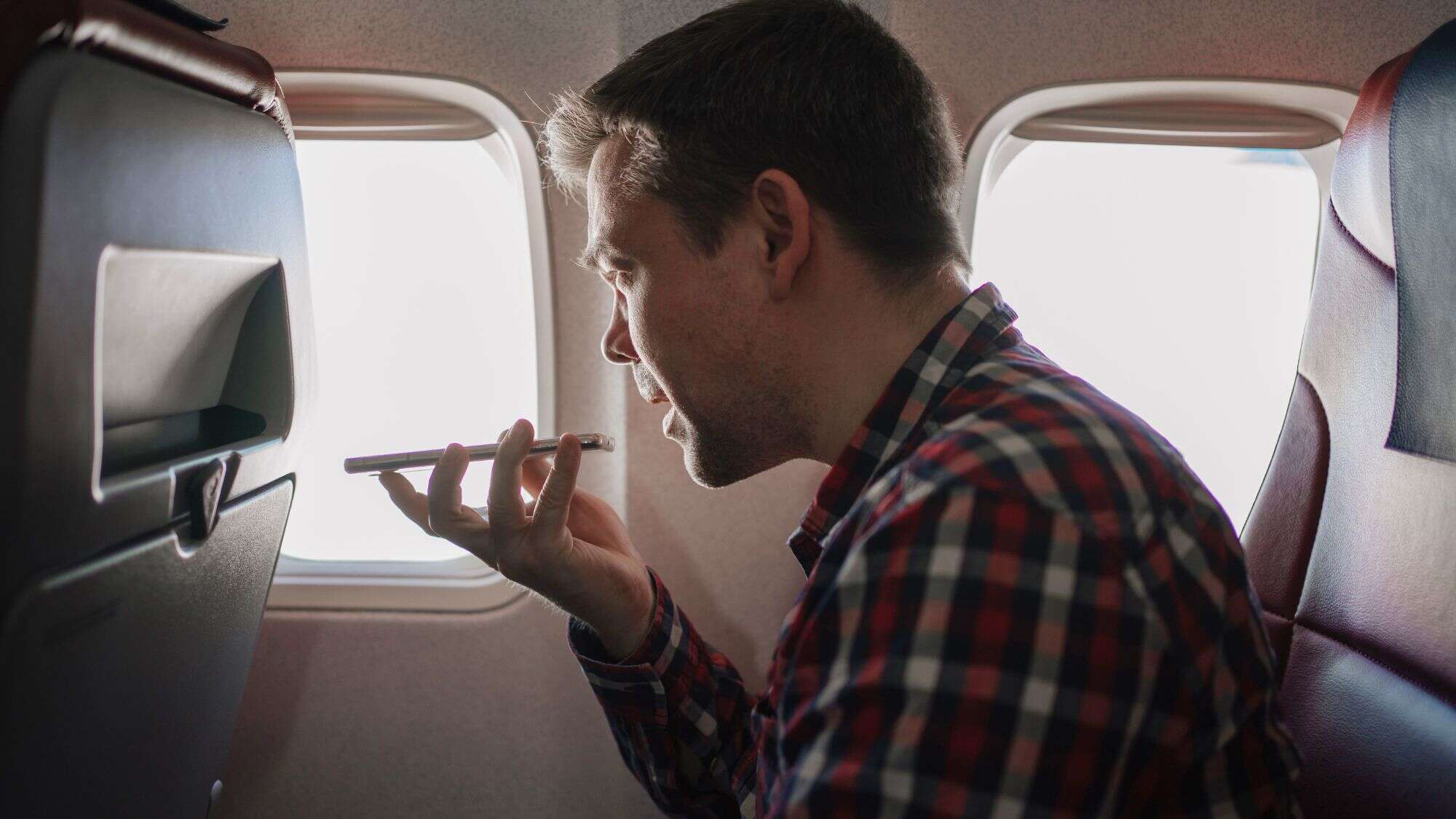 A man in a plaid shirt seated on an airplane speaks into his phone using the speakerphone feature, with the window and seatback in view.