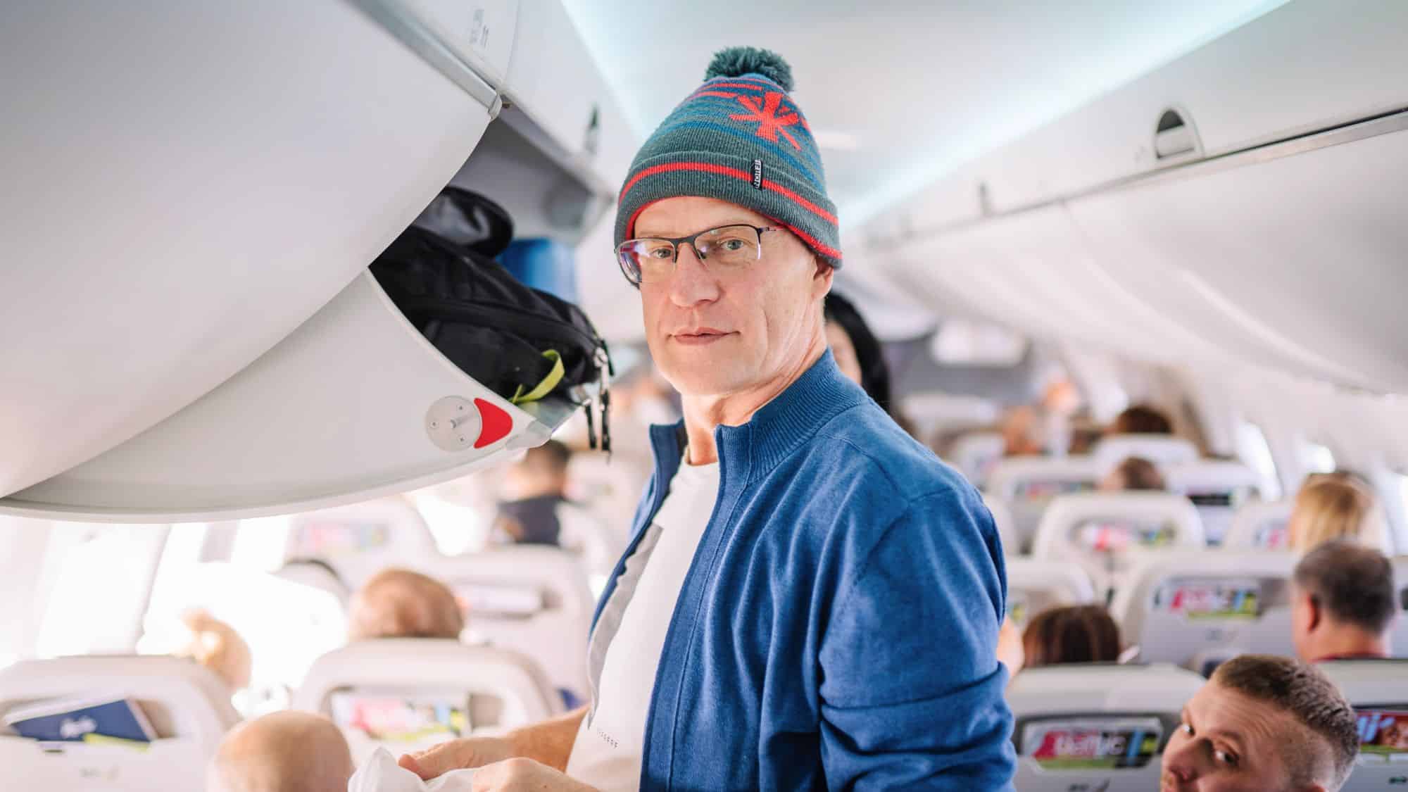 A man wearing glasses, a winter hat, and a blue jacket stands in the airplane aisle near the overhead compartment, facing the camera.
