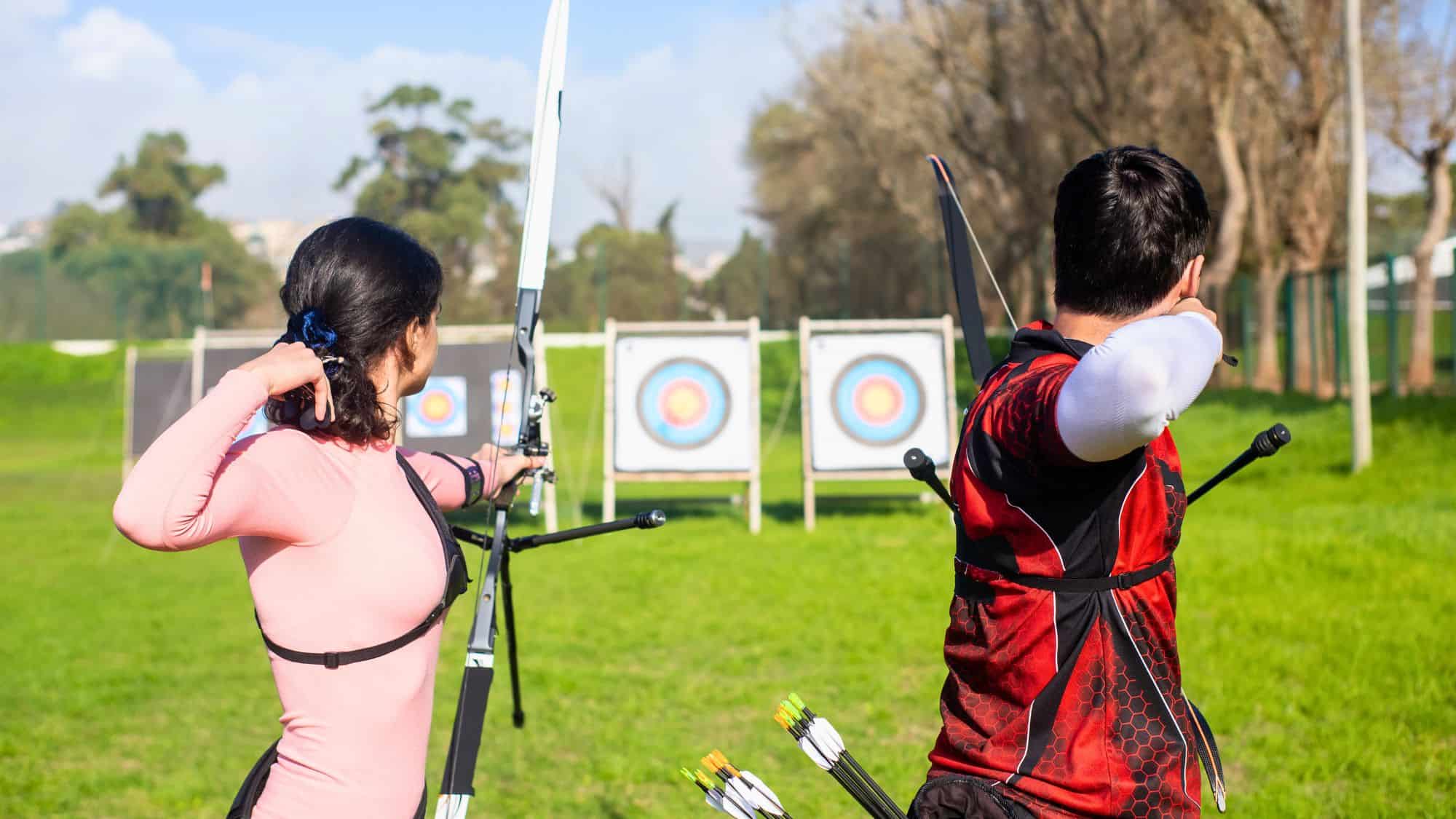 A man and a woman stand side by side at an outdoor archery range, drawing their bows with focused expressions. Targets with colorful bullseyes are set up in the background on a grassy field, framed by trees.