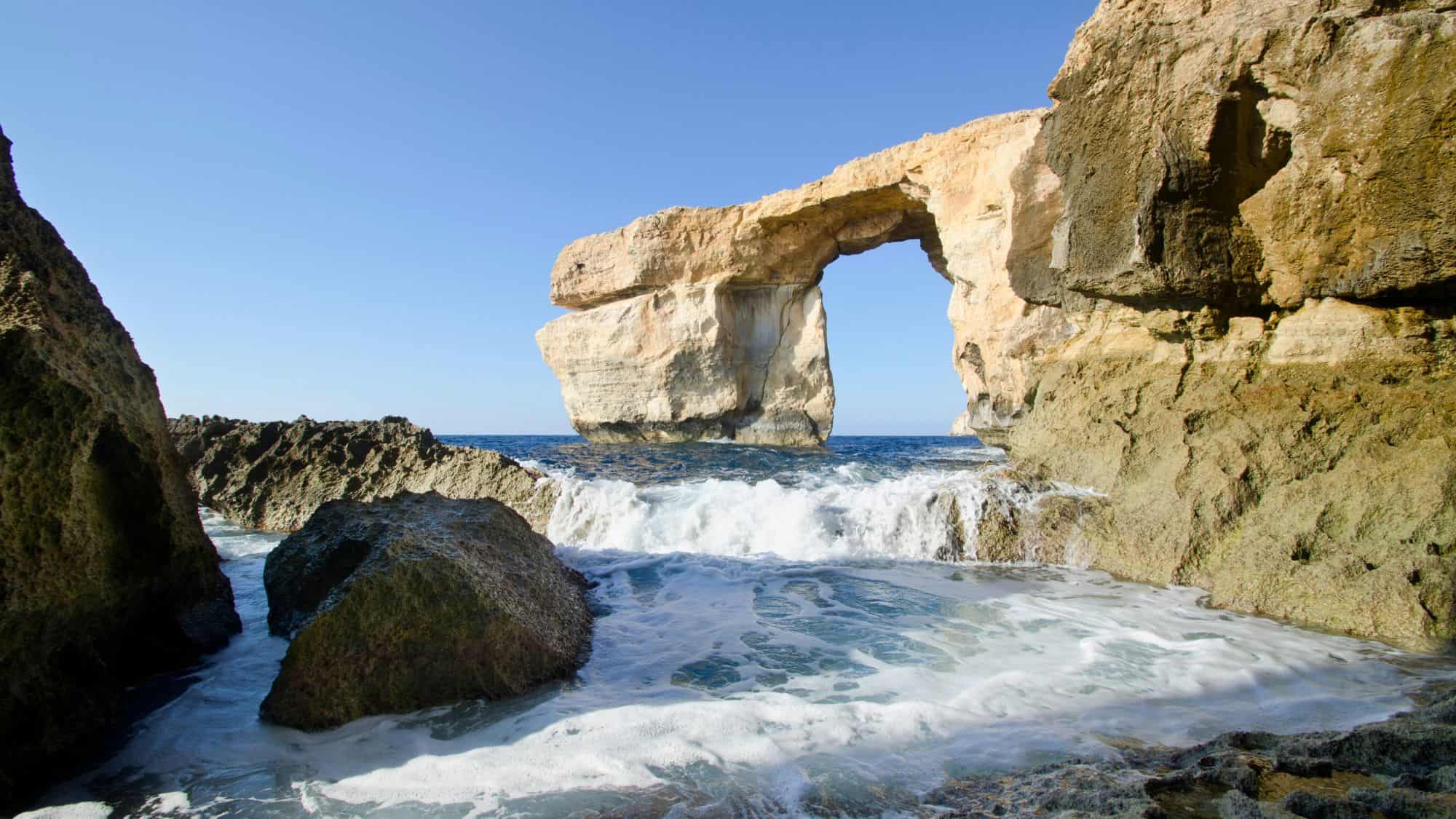 A natural limestone arch known as the Azure Window rises from the rocky coastline of Gozo, Malta, with waves crashing beneath and a clear blue sky overhead. The rugged textures of the surrounding cliffs frame the iconic sea arch.