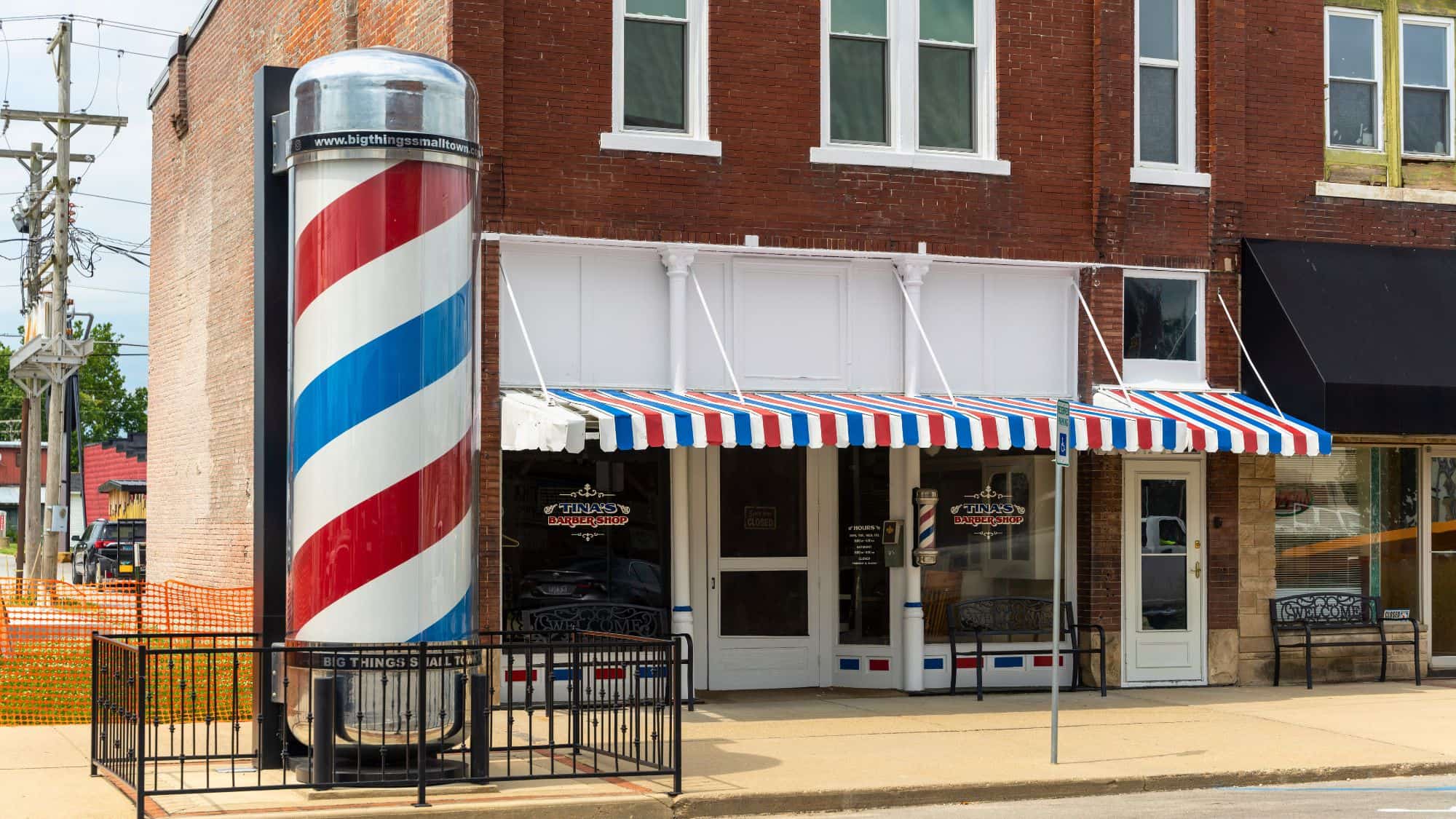 A giant barbershop pole stands outside a brick building with a red, white, and blue striped awning. The storefront, named "Tina’s Barbershop," has a traditional barber's pole beside the entrance, complementing the oversized decorative feature.