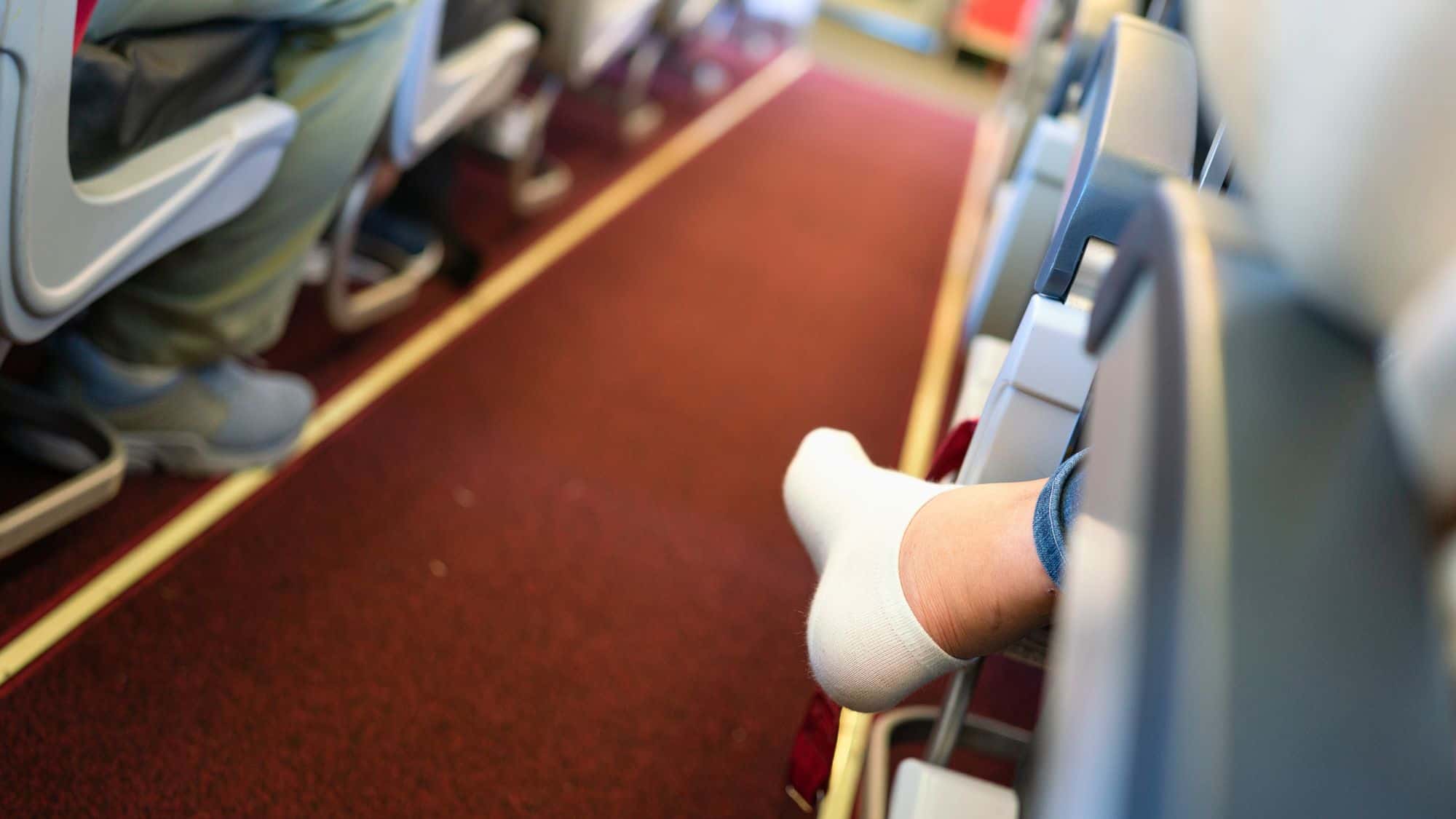 A passenger’s foot in a white sock rests against the aisle wall of an airplane, with red carpeting and nearby seated passengers in view.