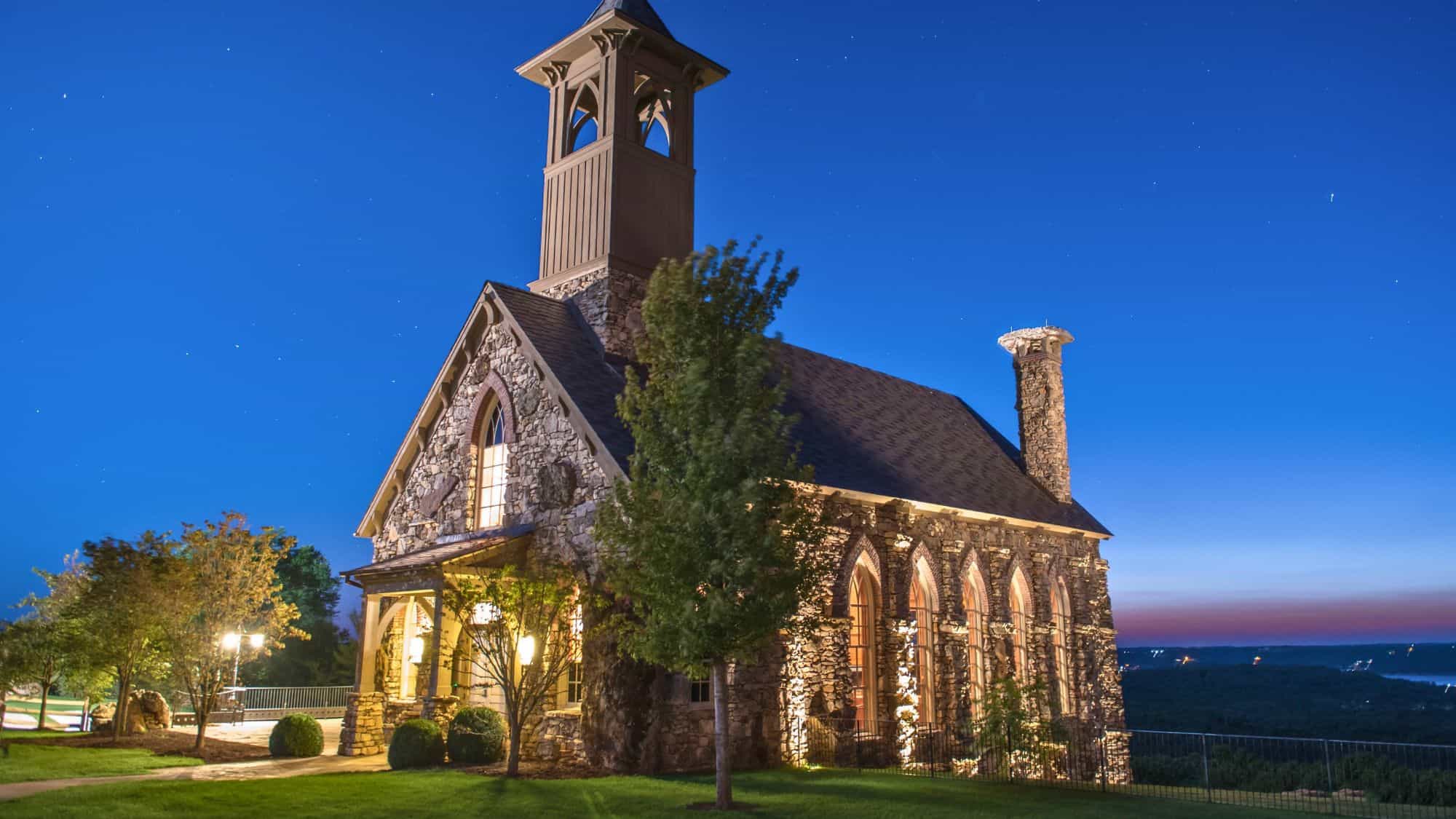 A beautifully lit stone chapel with arched windows and a bell tower stands against a deep blue evening sky. The surrounding landscape overlooks rolling hills and a distant body of water, creating a serene and picturesque setting.