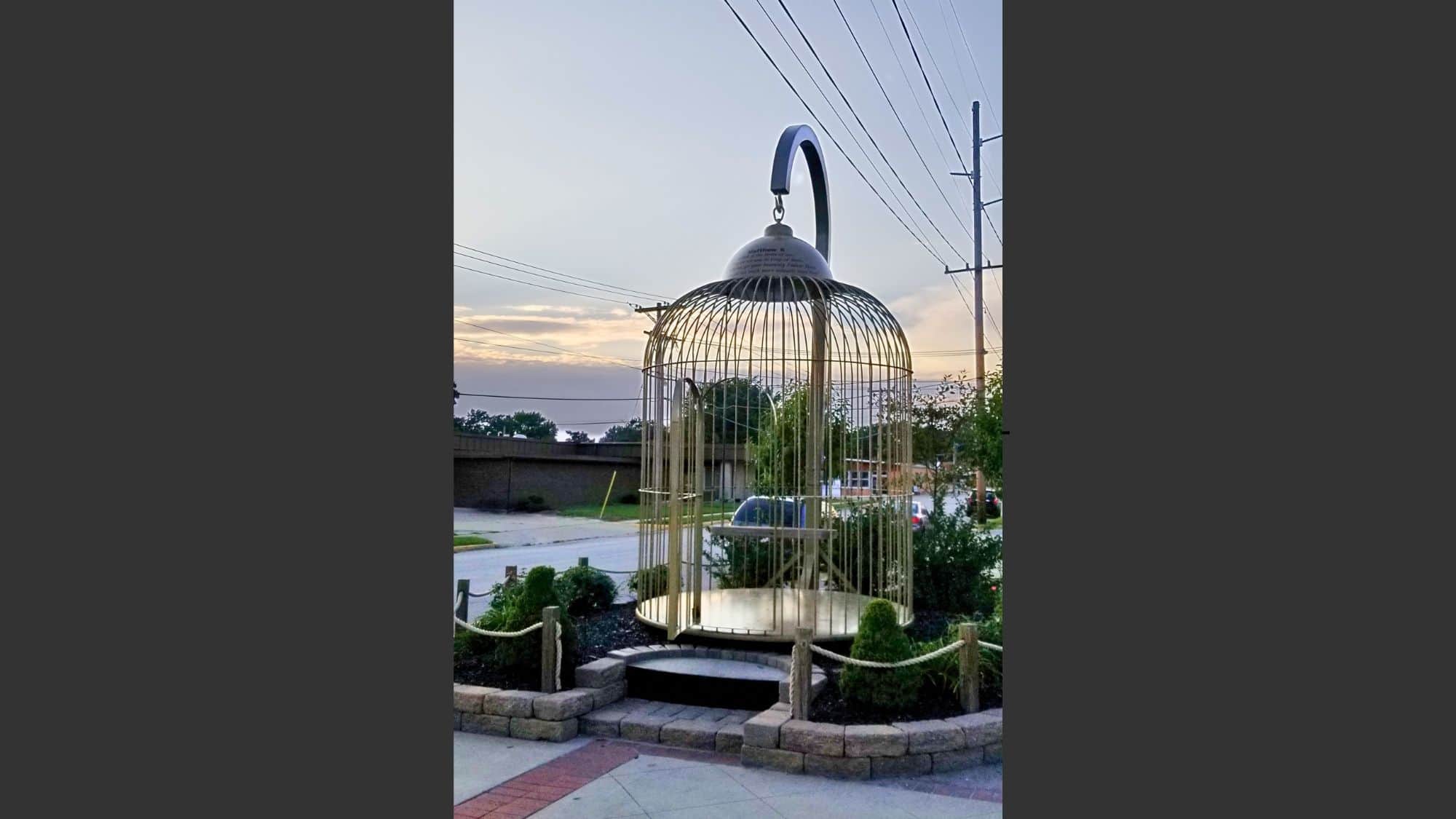 A massive golden birdcage sculpture sits in a landscaped area with a rope barrier and stone base. The open-air design and large handle on top give it the appearance of a whimsical, oversized birdcage set against a backdrop of utility poles and a sunset sky.