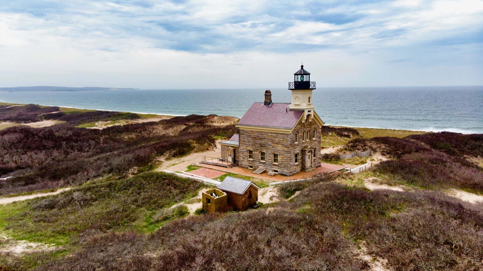 A historic lighthouse stands alone among rolling dunes, overlooking the vast ocean. The stone structure is surrounded by sandy pathways, with a dramatic sky above adding to the coastal charm.