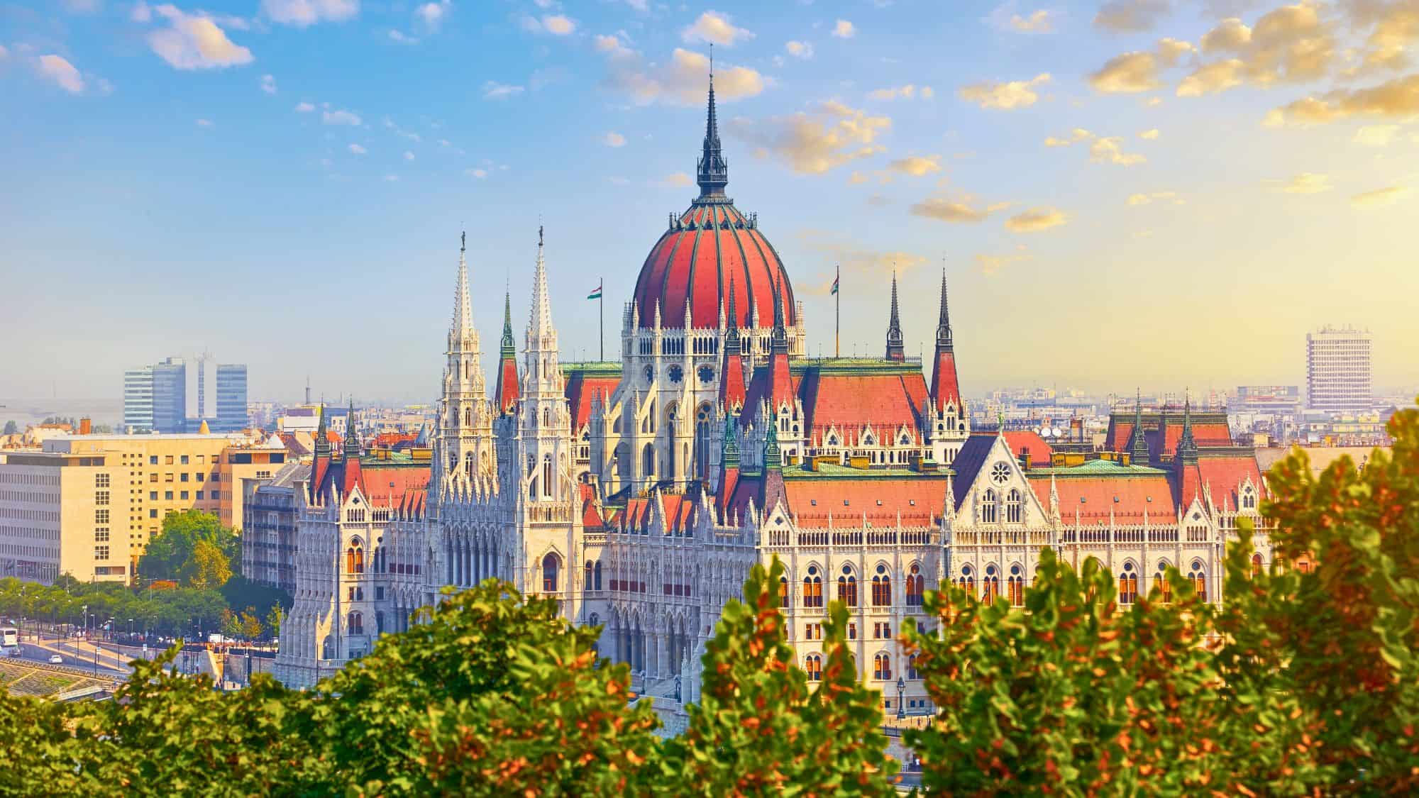 A breathtaking view of the Hungarian Parliament Building in Budapest, showcasing its neo-Gothic architecture with red-tiled rooftops and intricate spires. The golden light of sunrise highlights the stunning details.