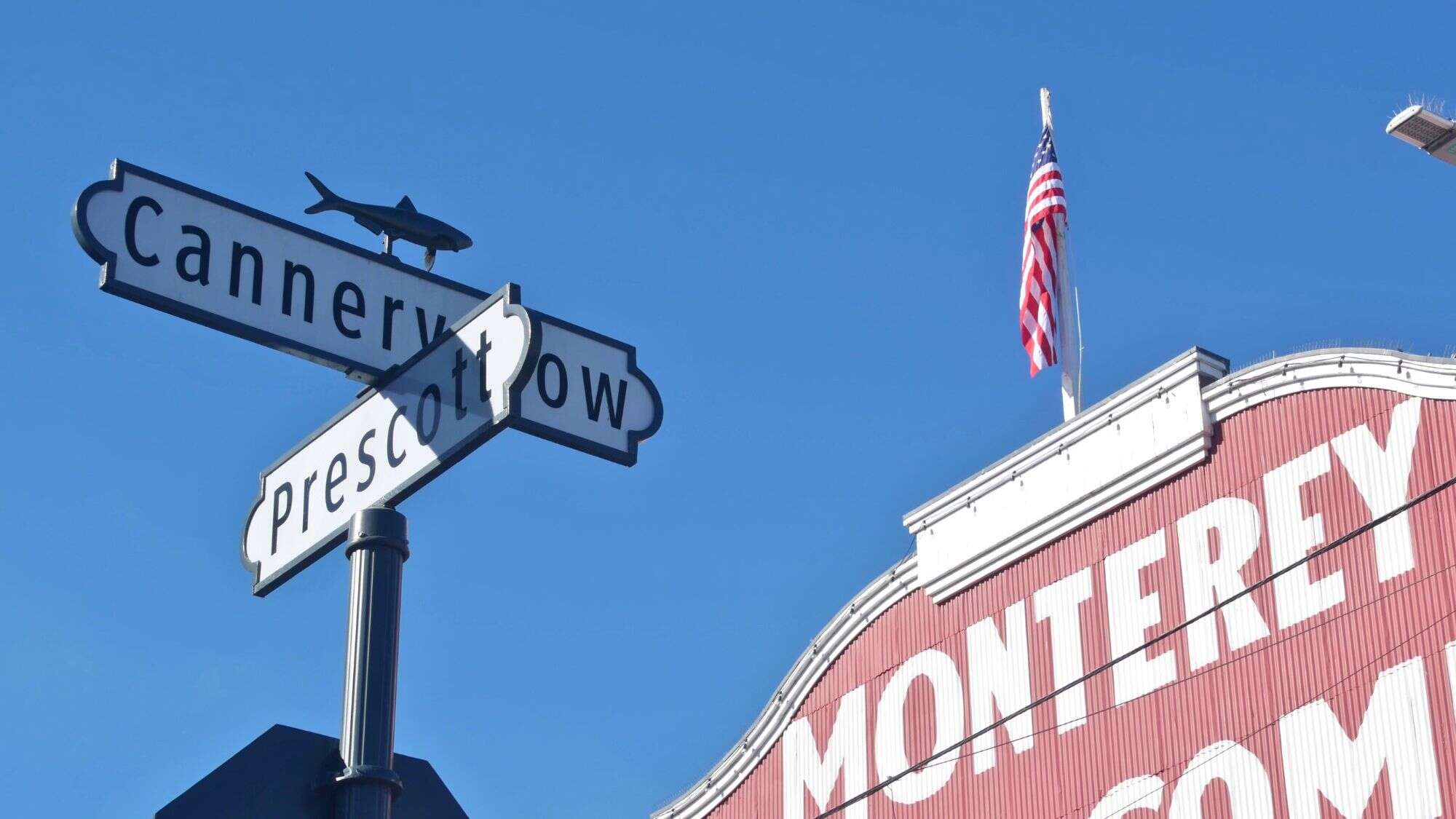A street sign at the intersection of Cannery Row and Prescott in Monterey, California, with a small fish figure perched on top. In the background, part of a red building with large white lettering spelling "MONTEREY" is visible, along with an American flag waving against a bright blue sky.