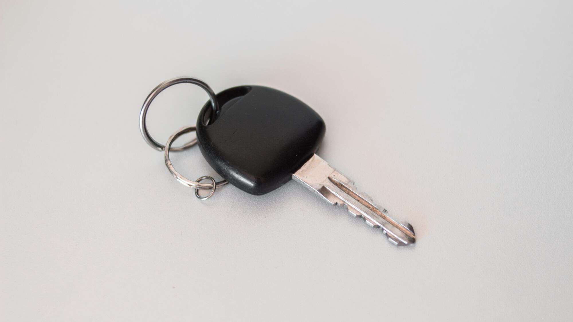 A single car key with a black plastic head is attached to two silver key rings, resting on a plain, light gray surface.