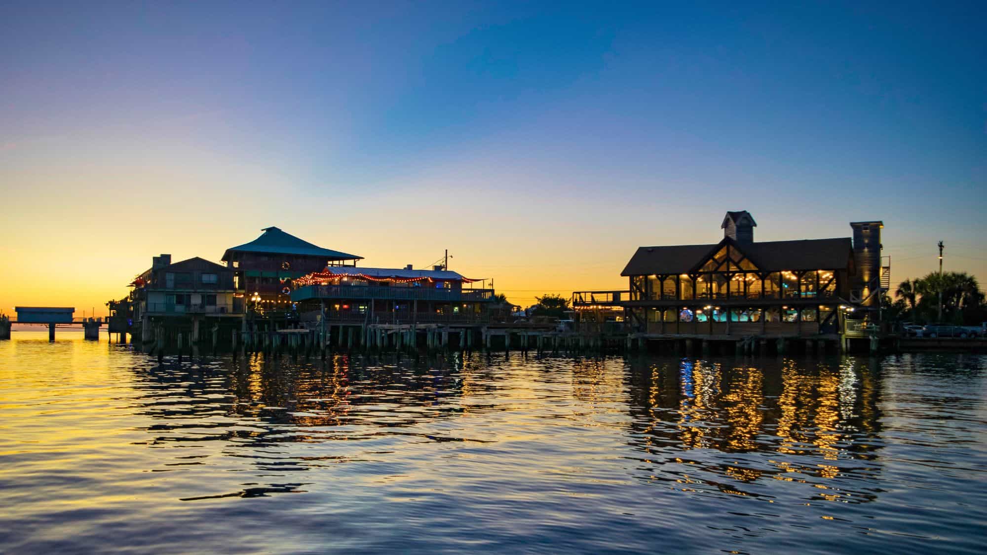 Charming waterfront restaurants in Cedar Key, Florida glow with string lights and warm interior lighting, reflecting beautifully off the calm bay as the sun sets behind them.