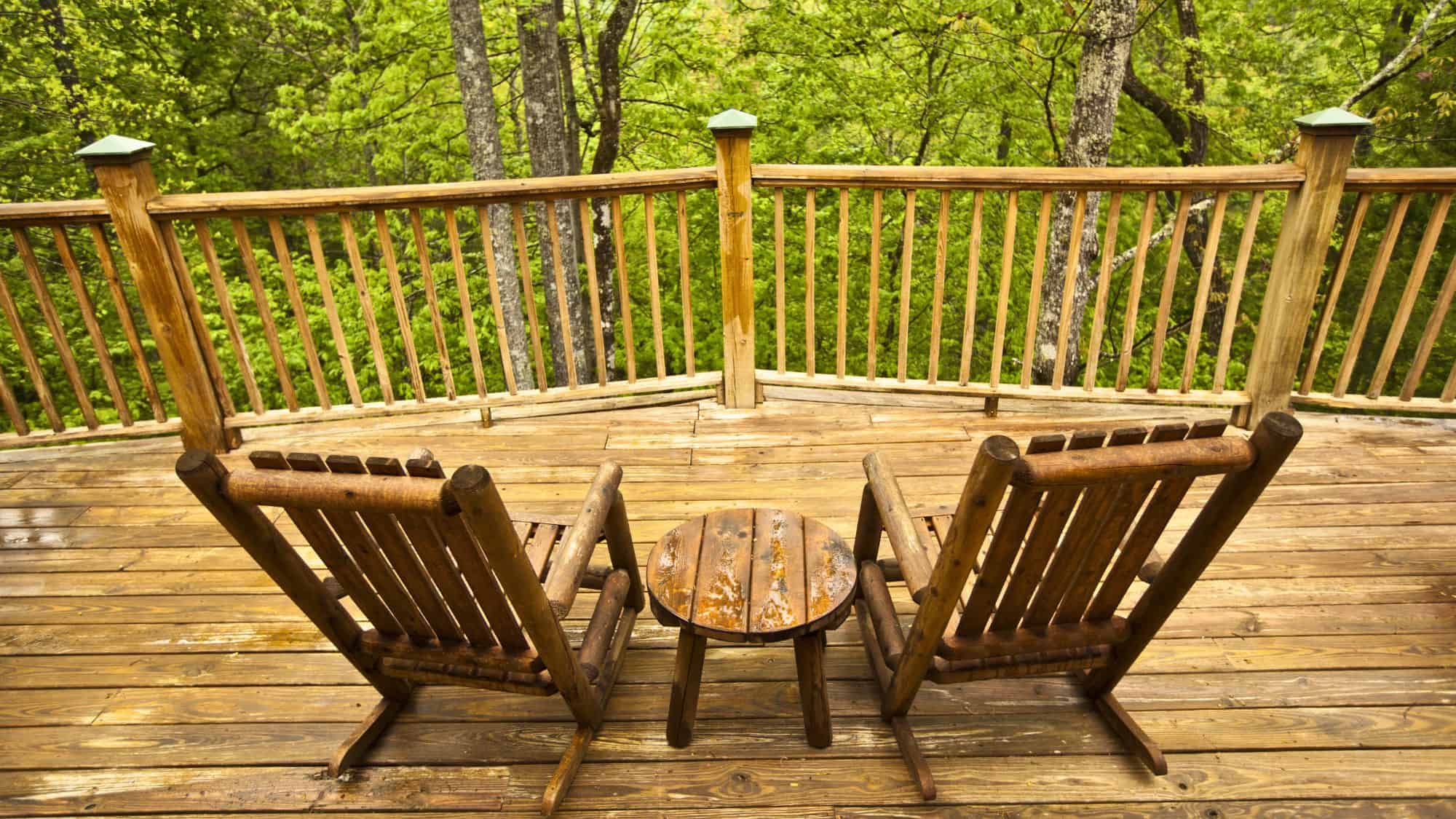 A rustic wooden deck with two Adirondack-style rocking chairs and a small round table, surrounded by a lush green forest. The wood appears slightly damp, enhancing the natural ambiance.