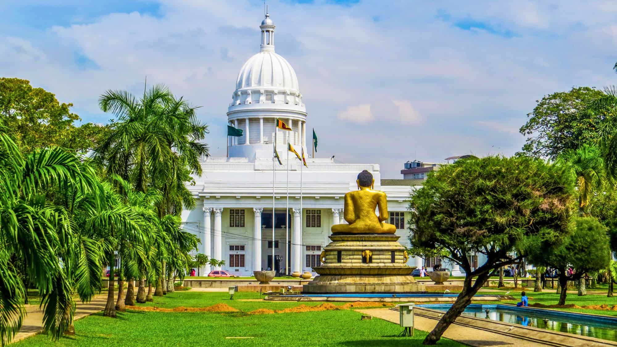A grand white-domed colonial-style building stands in the background, with a golden Buddha statue sitting in a meditative pose in the foreground. The lush green park surrounding the structure is dotted with palm trees, reflecting Colombo’s mix of history and spirituality.