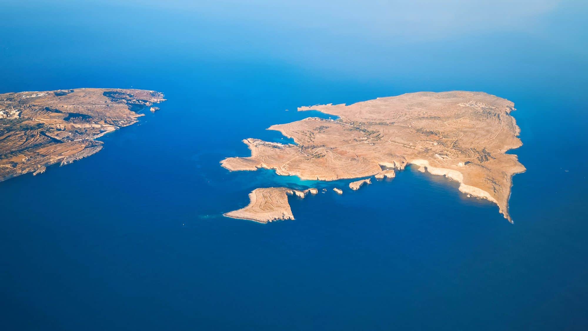 An aerial view of Comino Island, nestled between Malta and Gozo, showcases its arid, rugged terrain and jagged coastline surrounded by deep blue Mediterranean waters. The clarity of the sea highlights the shallow coves and inlets around the island.