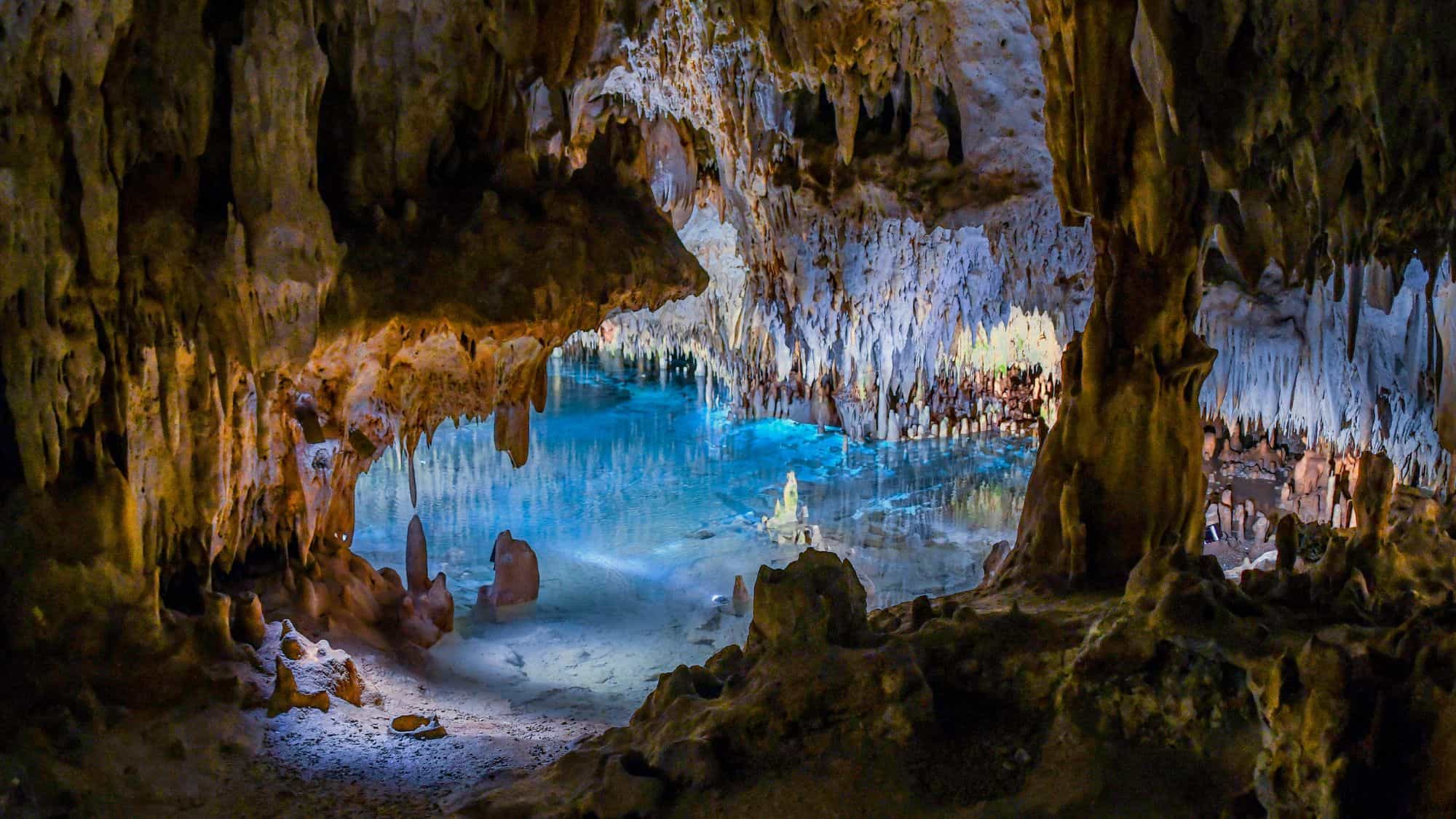 A stunning underground cave filled with dramatic stalactites and stalagmites, illuminated by soft lighting. A crystal-clear underground pool reflects the rock formations, creating a surreal, otherworldly atmosphere.