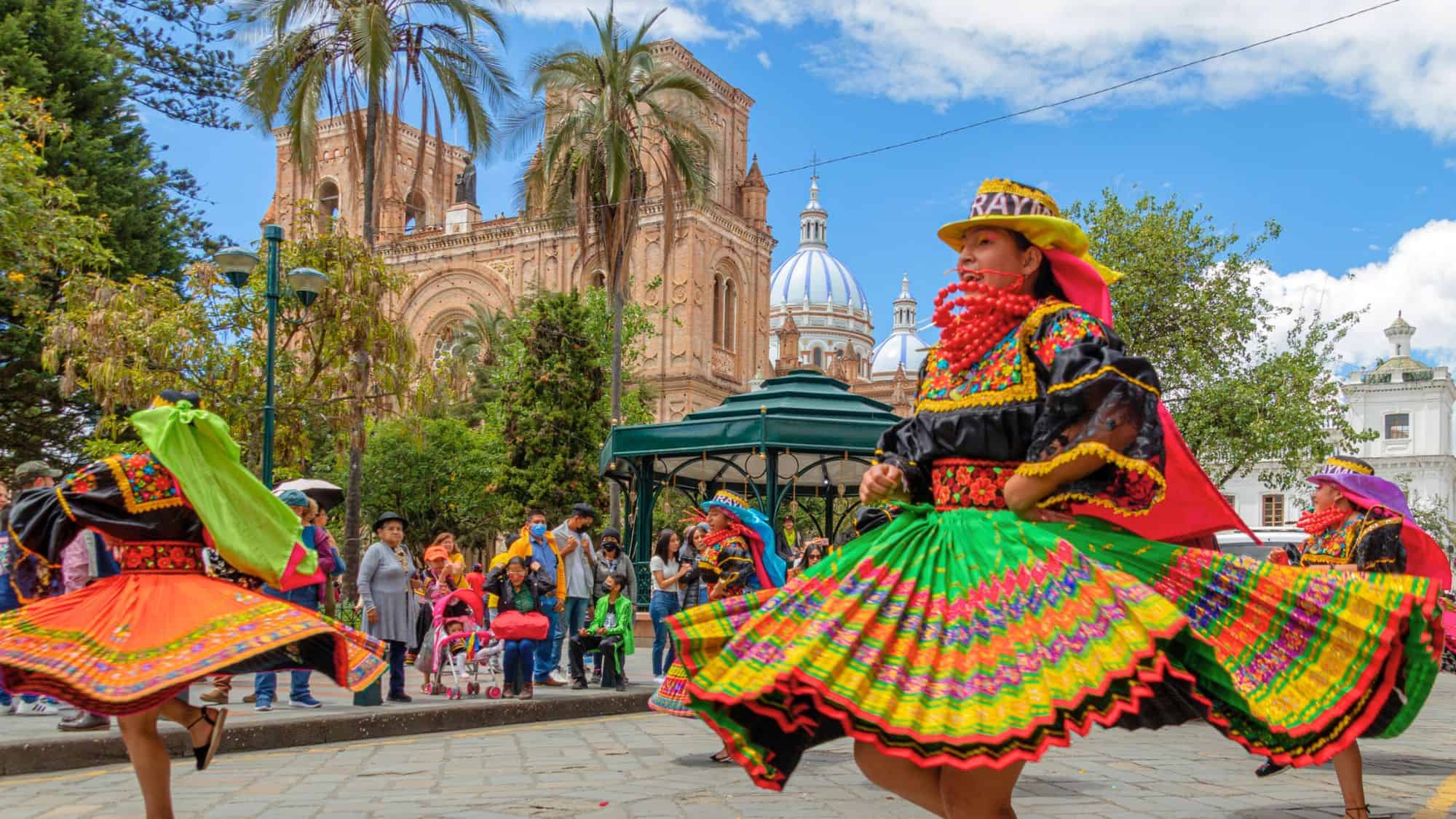 Women dressed in vibrant traditional Andean attire twirl in a folkloric dance, their colorful skirts fanning out in motion. Behind them, the iconic blue-domed Cathedral of the Immaculate Conception towers over a lively plaza filled with spectators.