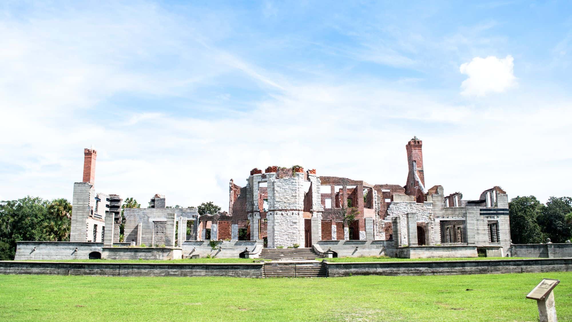 The ruins of a grand estate sit against a backdrop of blue sky, their weathered stone walls and towering chimneys remnants of a once-majestic mansion. Lush green grass surrounds the site, evoking a sense of history and mystery.