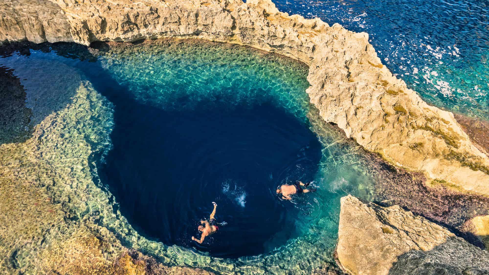 Two snorkelers swim inside the Deep Blue Hole near Gozo, a circular, dark blue natural sea pool surrounded by jagged limestone rock and crystal-clear shallows. The intense color contrast between the hole and the surrounding water is striking.