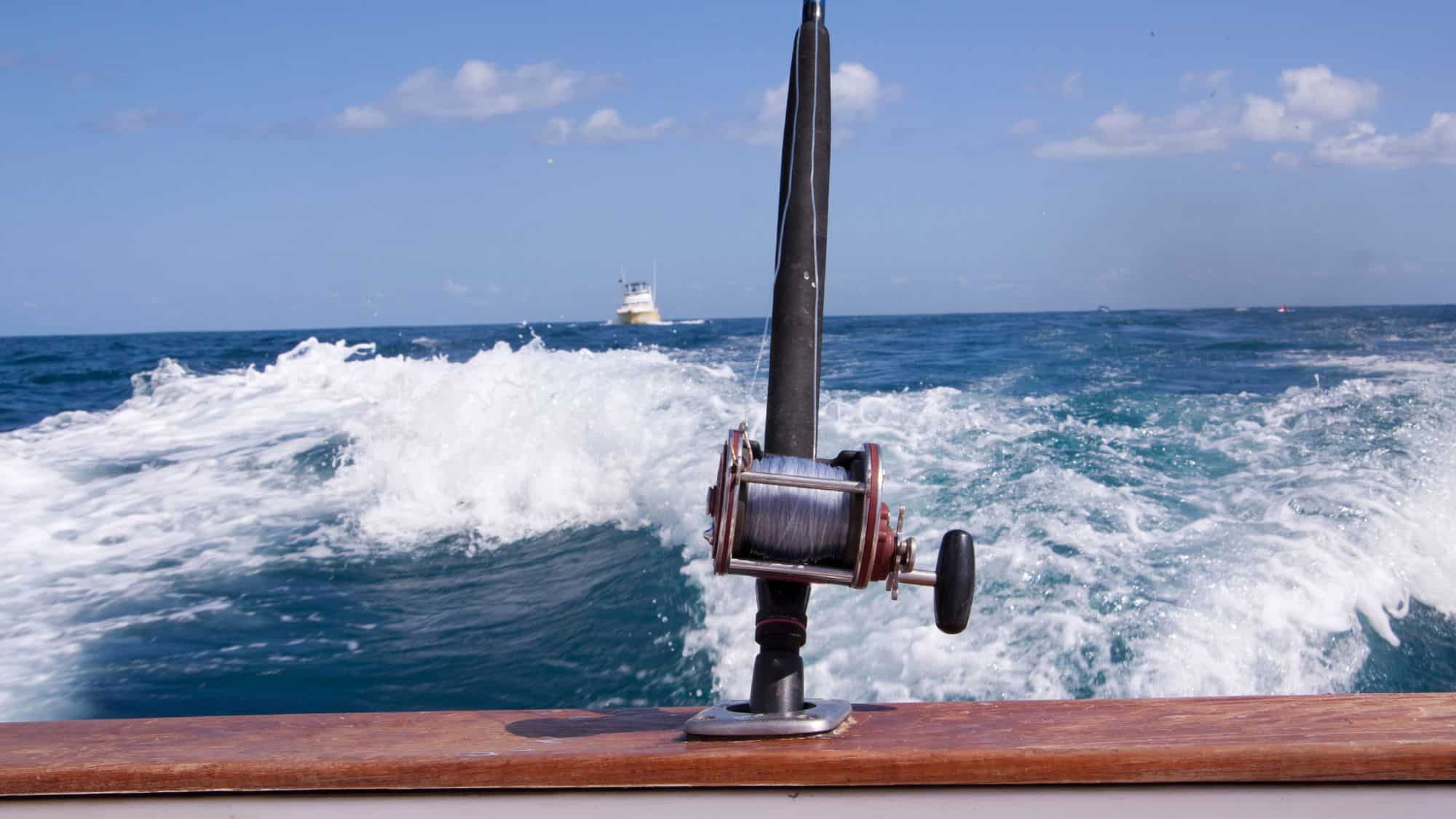 A fishing rod is mounted on the back of a boat, cutting through the deep blue ocean, leaving a foamy white wake. A fishing boat is visible in the distance under a bright sky, capturing the excitement of offshore fishing.