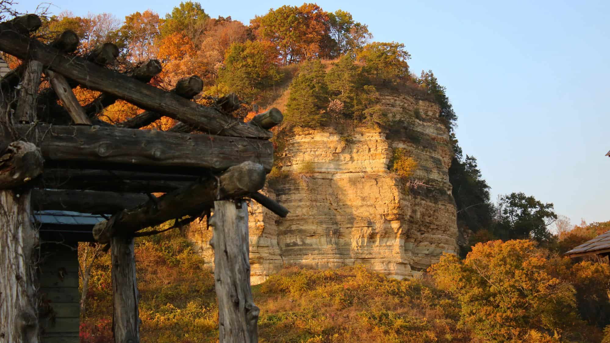 A sunlit limestone bluff covered in fall foliage rises behind a rustic wooden structure, showcasing the rugged beauty of the Driftless Area in autumn.
