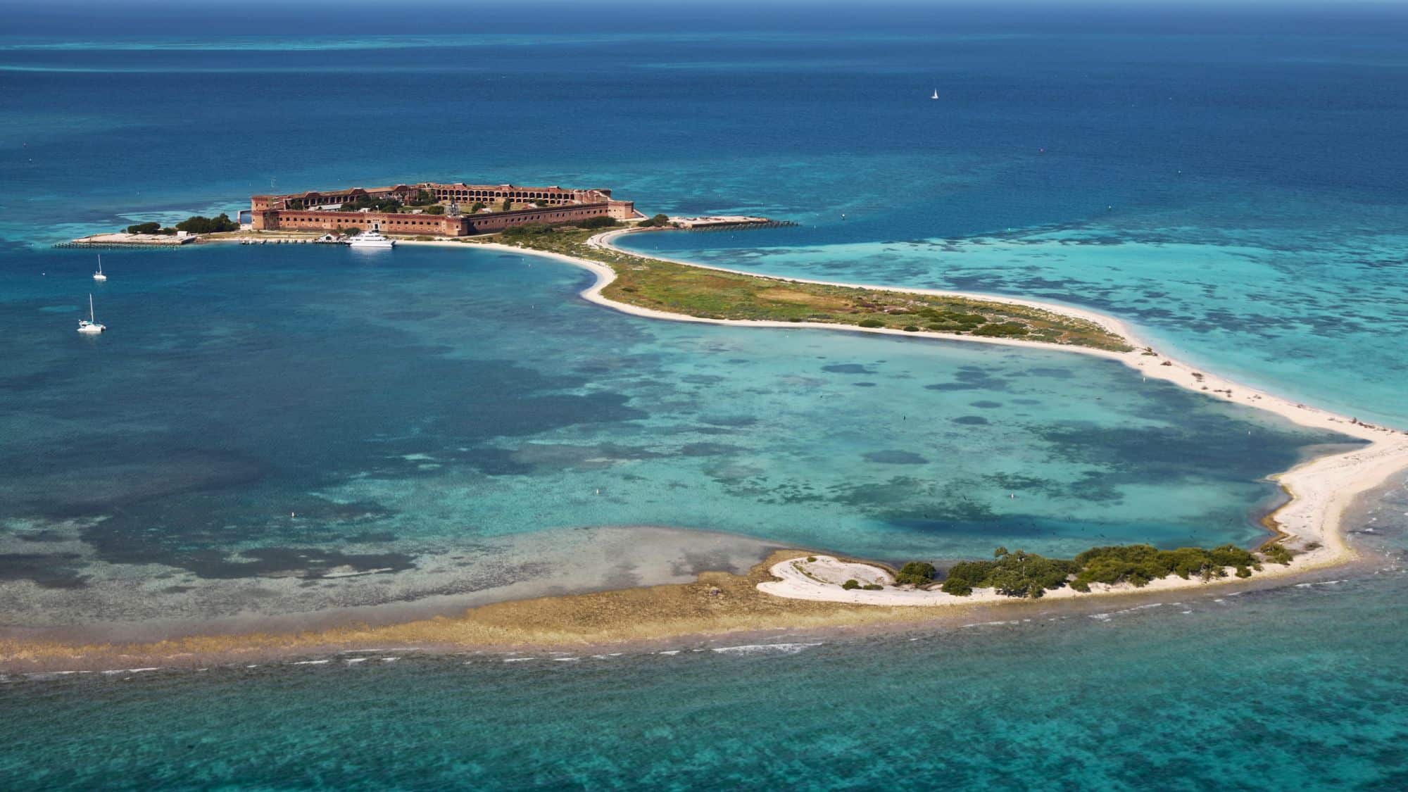 An aerial view of a remote island fortress, surrounded by turquoise waters and coral reefs. The massive brick structure of Fort Jefferson stands prominently on the sandy shoreline, with boats anchored nearby in the clear, shallow waters.