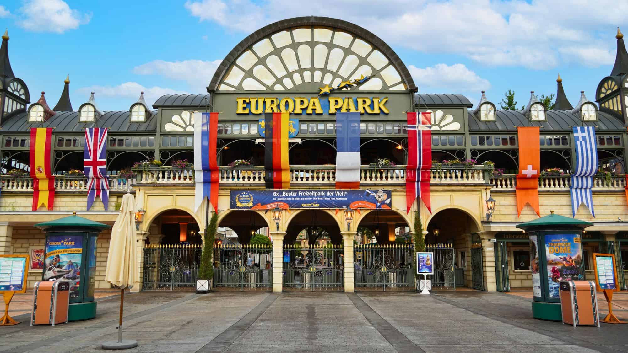 The grand entrance of Europa-Park in Germany, adorned with international flags and a large sign welcoming visitors. The arched glass and metal facade give it a classic European fairground look.