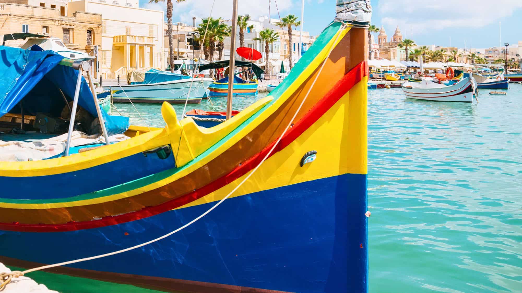 A close-up of a brightly painted traditional Maltese fishing boat, or luzzu, featuring the symbolic "Eye of Osiris" near the bow. The bold colors—yellow, red, blue, and green—pop against the turquoise harbor and coastal town backdrop.