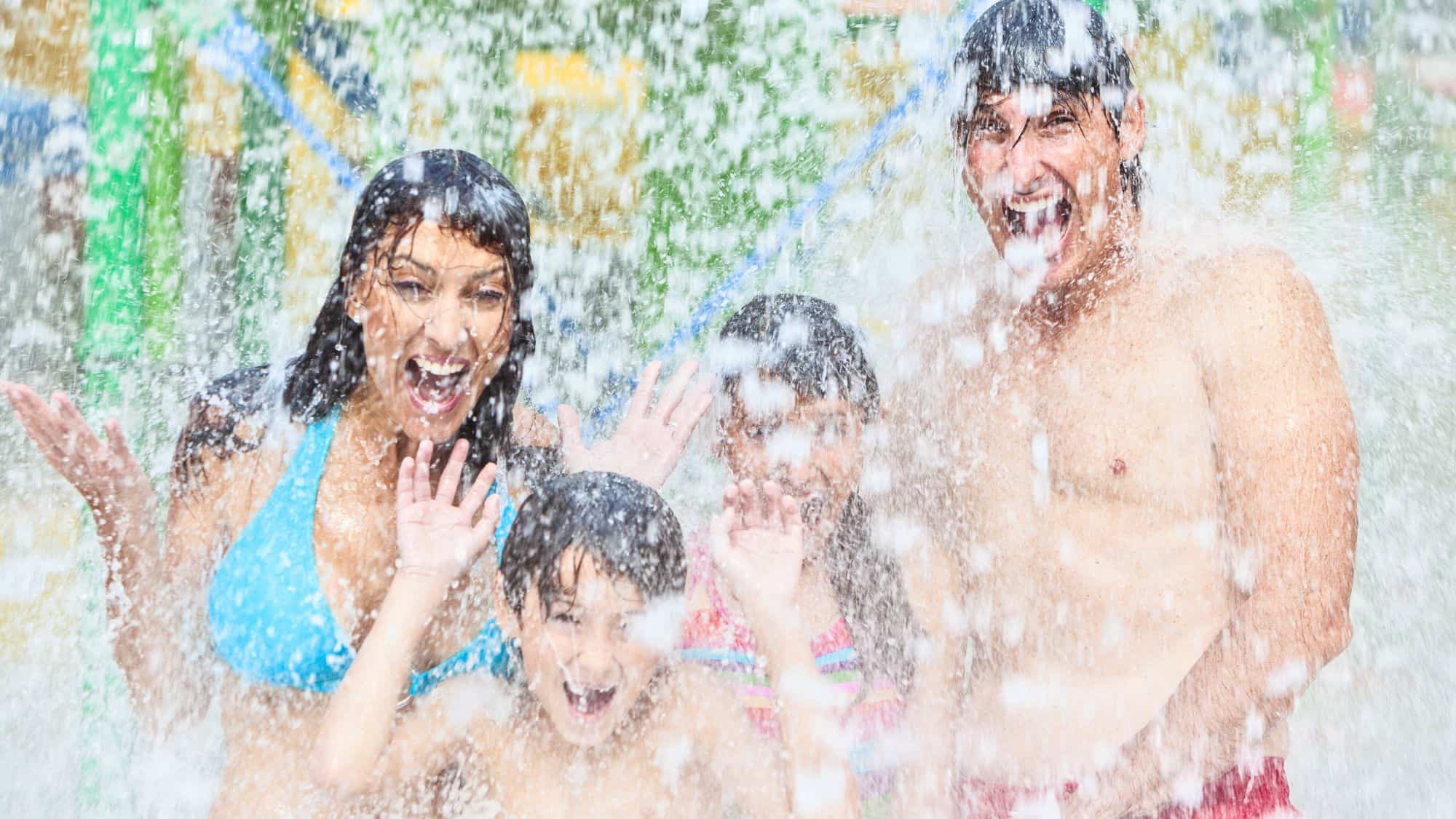 A joyful family of four, two parents and two children, laughing as they get drenched under a cascading waterfall at a water park. Their excitement and wide smiles capture the fun of a summer day.