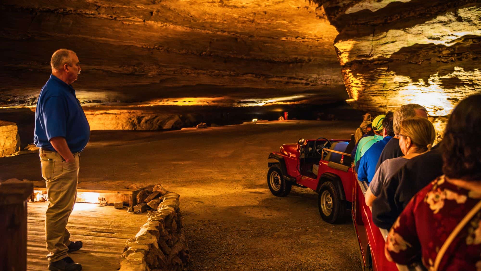 A guided tour inside Fantastic Caverns, where visitors ride in an open-air tram through the dimly lit cave, listening to a guide in a blue shirt explaining the geological formations surrounding them.