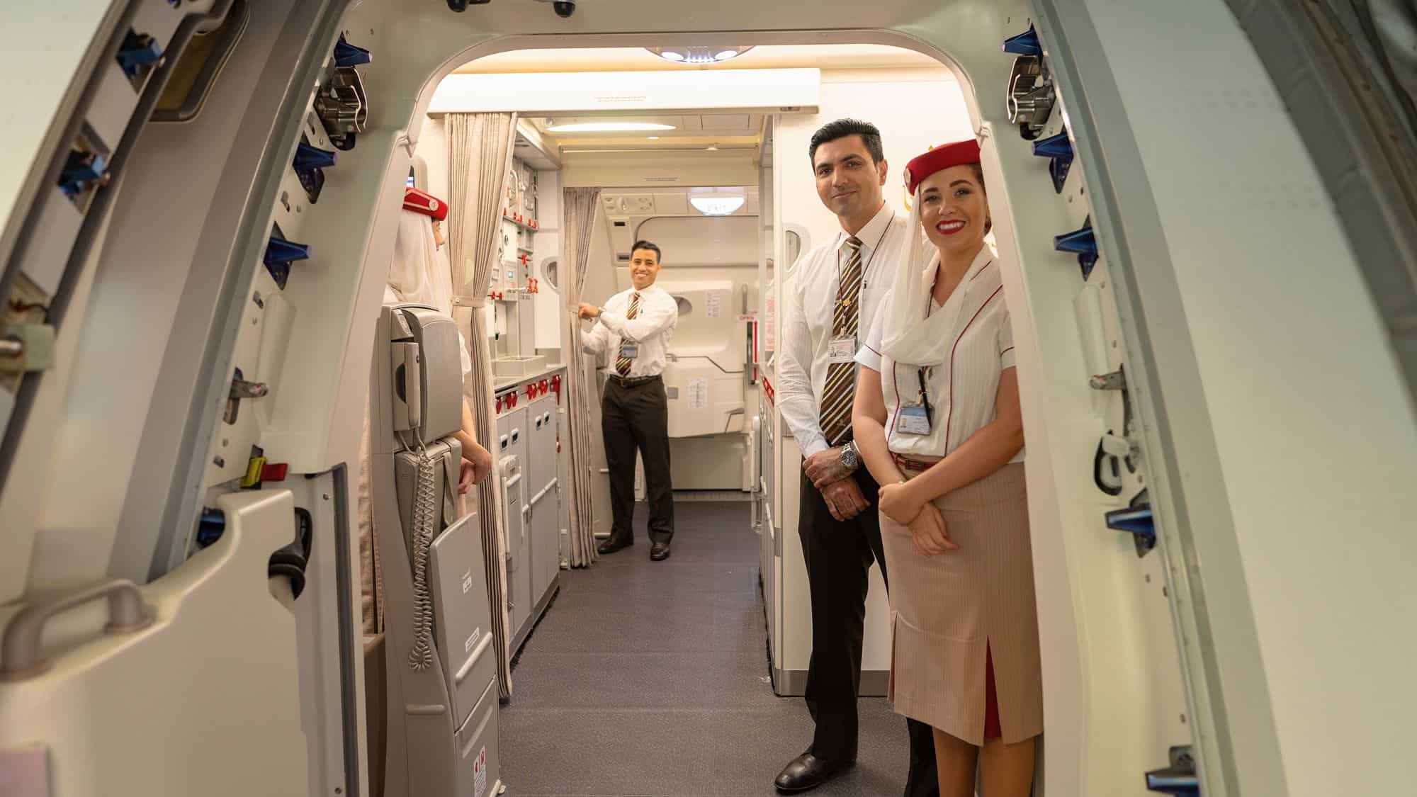 Three flight attendants stand smiling at the entrance of an airplane, welcoming passengers onboard, with one holding back a curtain to the cabin.