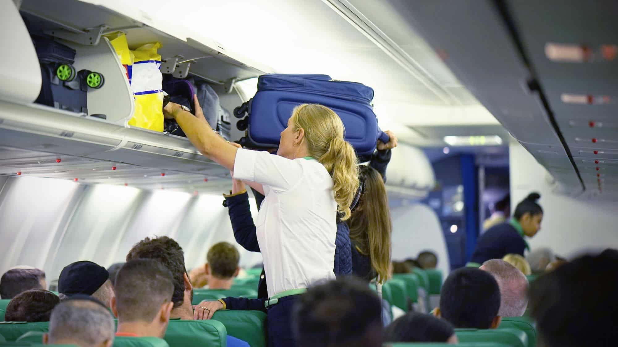 A blonde flight attendant assists passengers by placing luggage into an overhead bin on a crowded airplane filled with green seats.