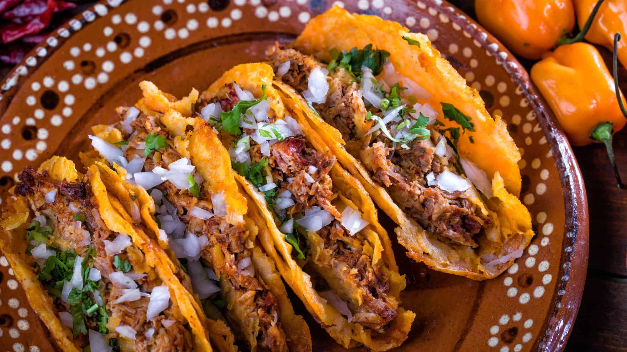A plate of crispy, golden frybread tacos filled with shredded, spiced meat and topped with fresh onions and cilantro. The tacos are served on a decorative ceramic plate, accompanied by spicy habanero peppers in the background.