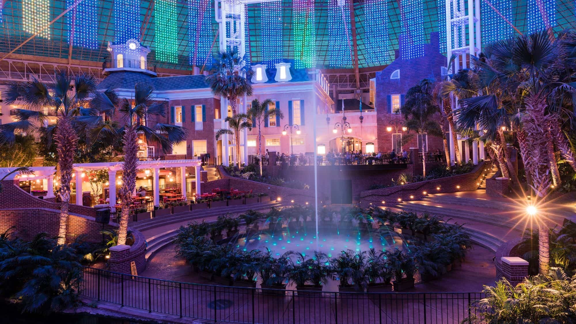 A beautifully illuminated atrium with colorful LED lights hanging from the ceiling, highlighting a grand fountain in the center. The scene is framed by palm trees, cozy outdoor dining, and a Southern-style facade.