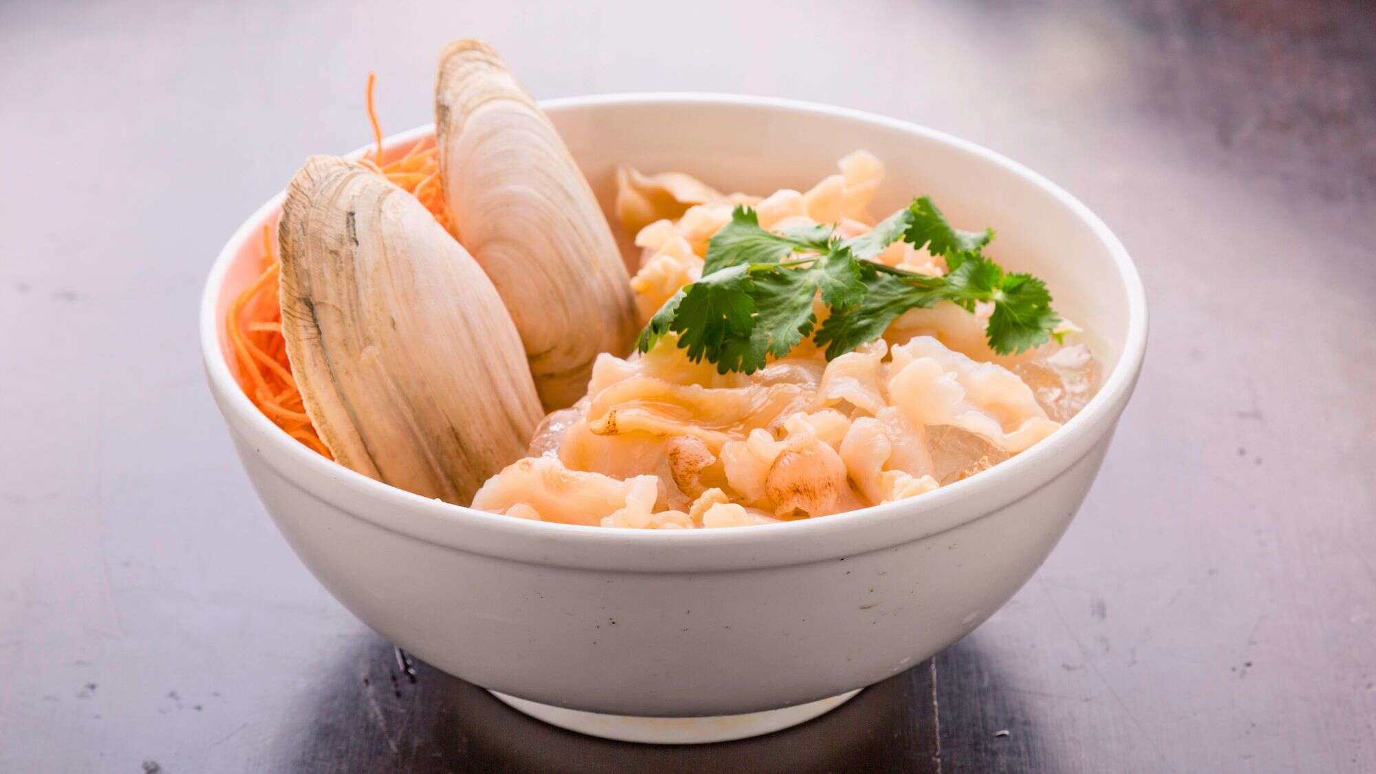 A white bowl filled with thinly sliced geoduck sashimi, arranged on ice with two large clamshells. Garnished with fresh cilantro and shredded carrots, the dish showcases a delicacy from the Pacific Northwest