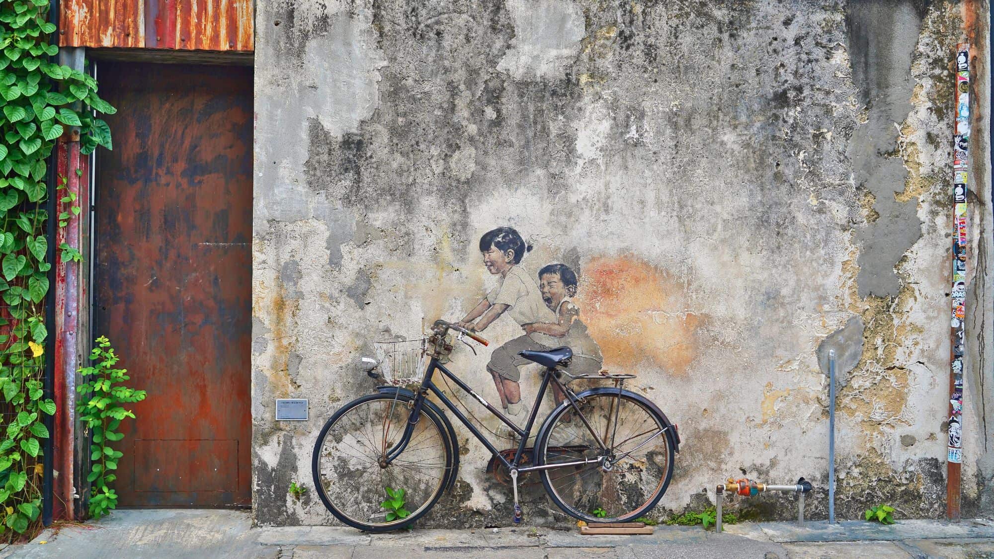 A famous street art mural on a weathered wall depicts two children riding a real bicycle, blending art with urban scenery. The textured wall, creeping green vines, and an old rusty door add to the historic charm of George Town’s UNESCO-listed heritage streets.