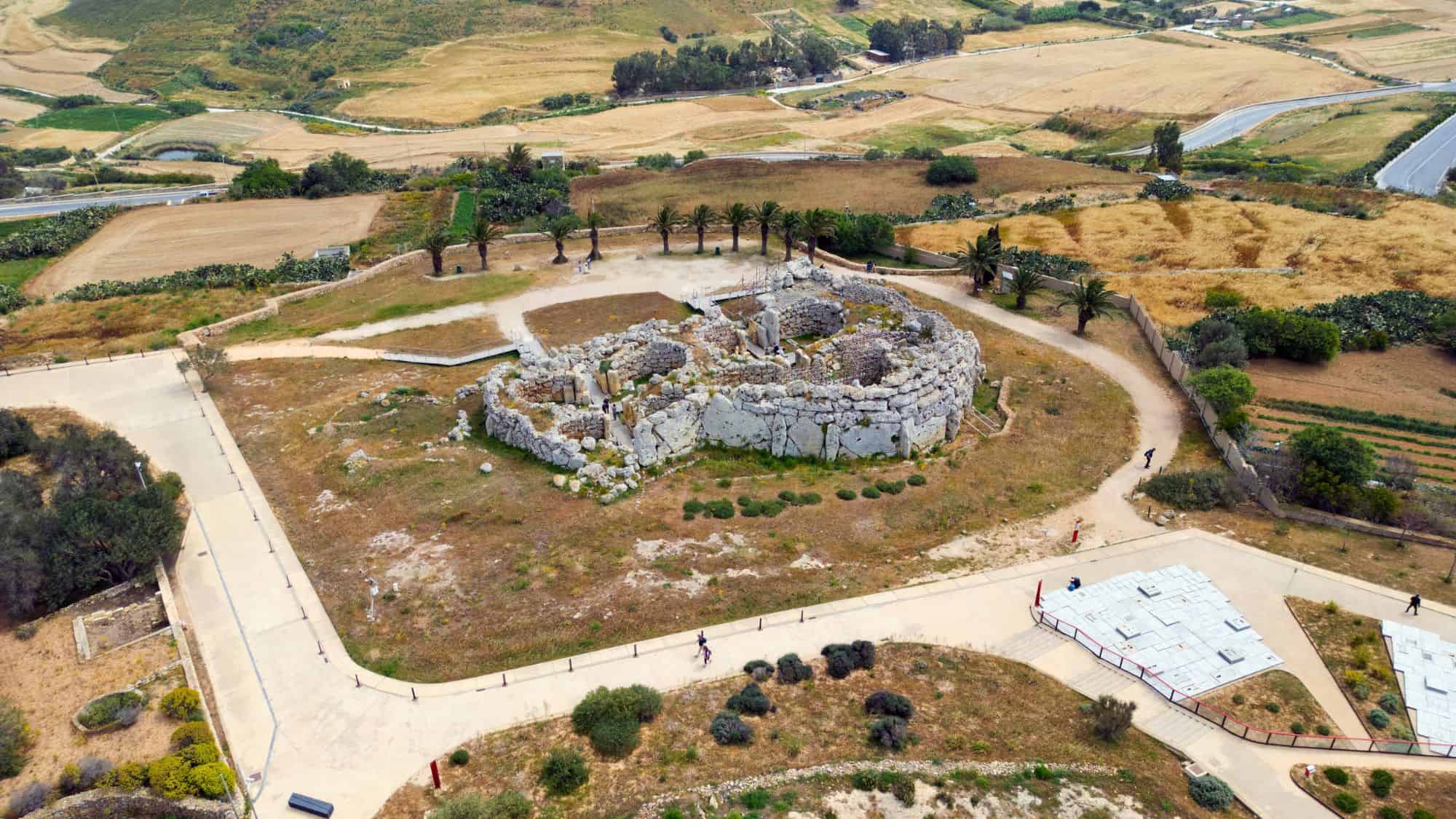 Aerial view of the ancient Ġgantija megalithic temples in Malta, showcasing massive stone ruins surrounded by footpaths, fields, and palm trees in the countryside.