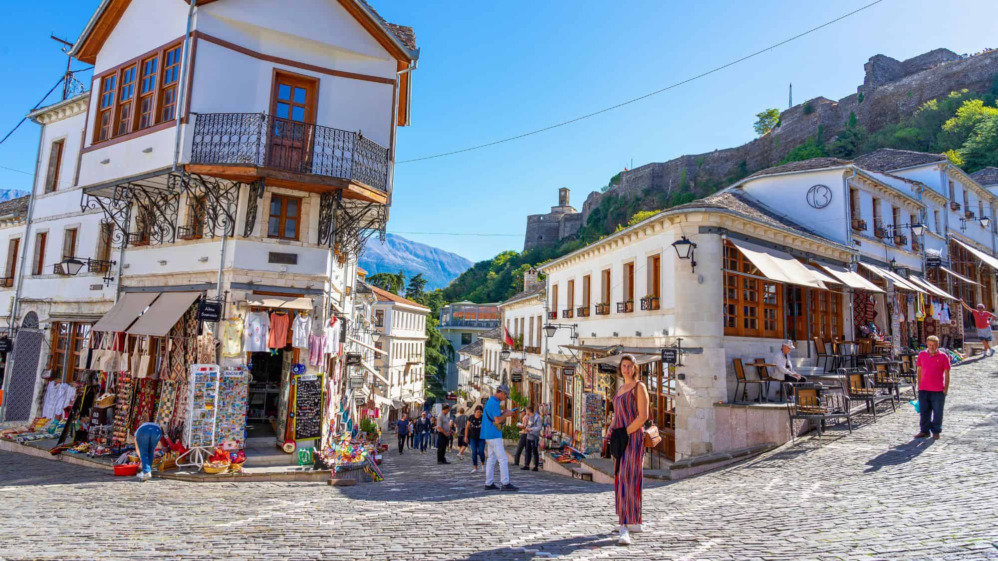 A cobblestone street lined with Ottoman-style white houses and bustling souvenir shops leads up to a historic castle in the background. A woman in a stylish outfit walks through the charming bazaar, surrounded by locals and tourists exploring the well-preserved old town.