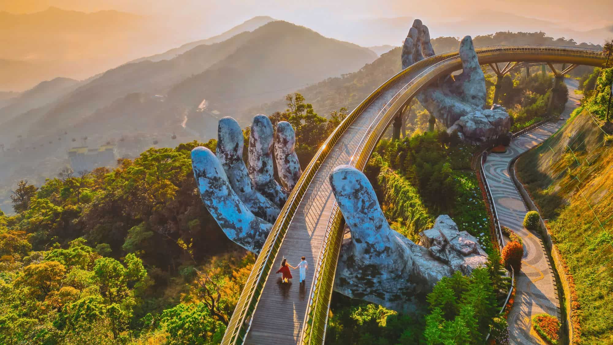 A stunning view of the Golden Bridge in Da Nang, Vietnam, held up by massive stone hands. The bridge curves gracefully over lush green mountains, with soft mist and golden sunrise light adding to the dreamlike atmosphere.