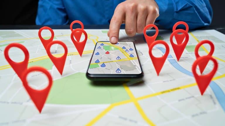 A person in a blue shirt interacts with a smartphone placed on a paper map, surrounded by multiple red location pins symbolizing various destinations. The image represents digital navigation and location-based services.