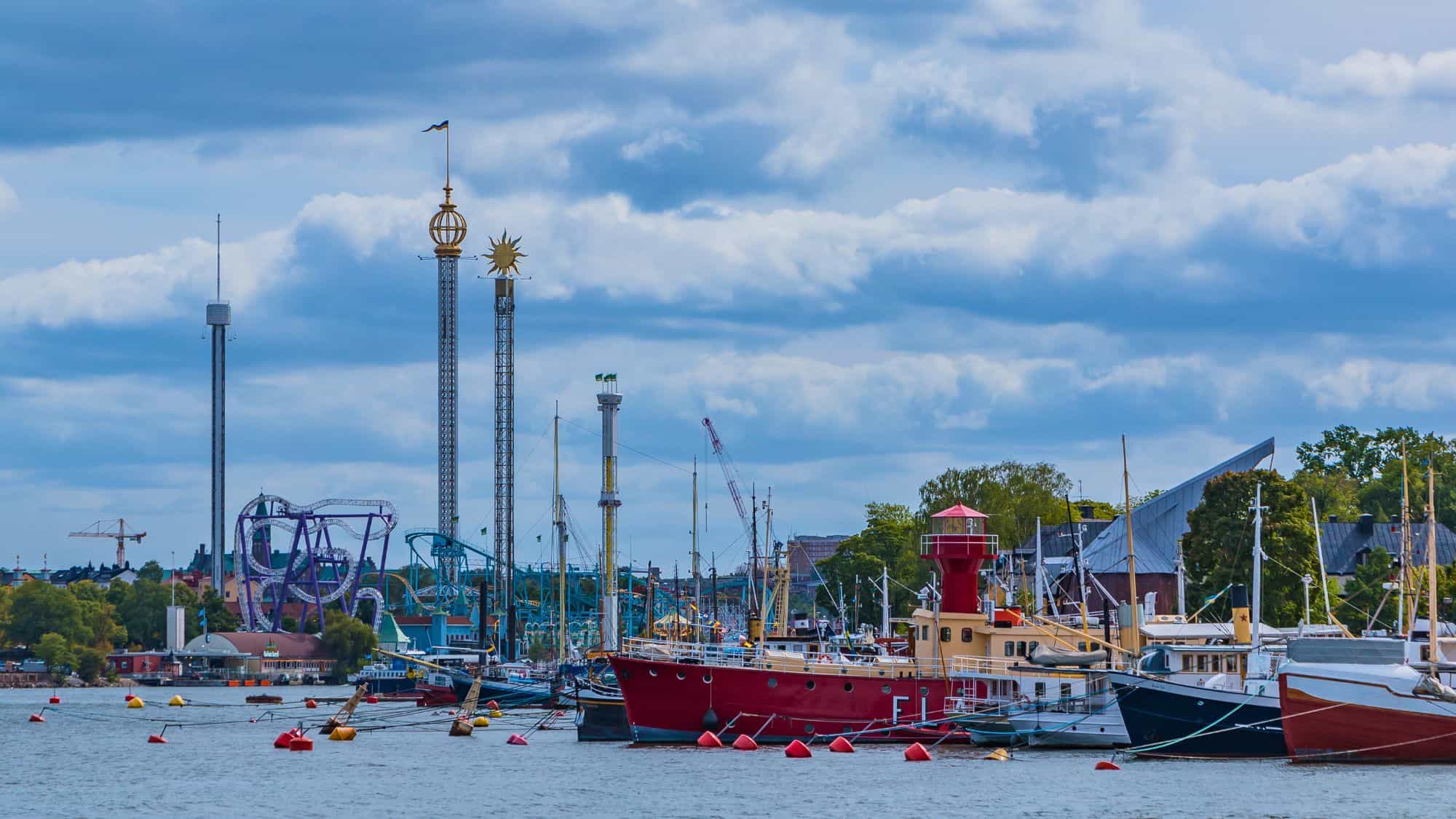 A scenic view of Gröna Lund amusement park in Stockholm, Sweden, with its towering rides and roller coasters visible beyond the waterfront filled with boats.