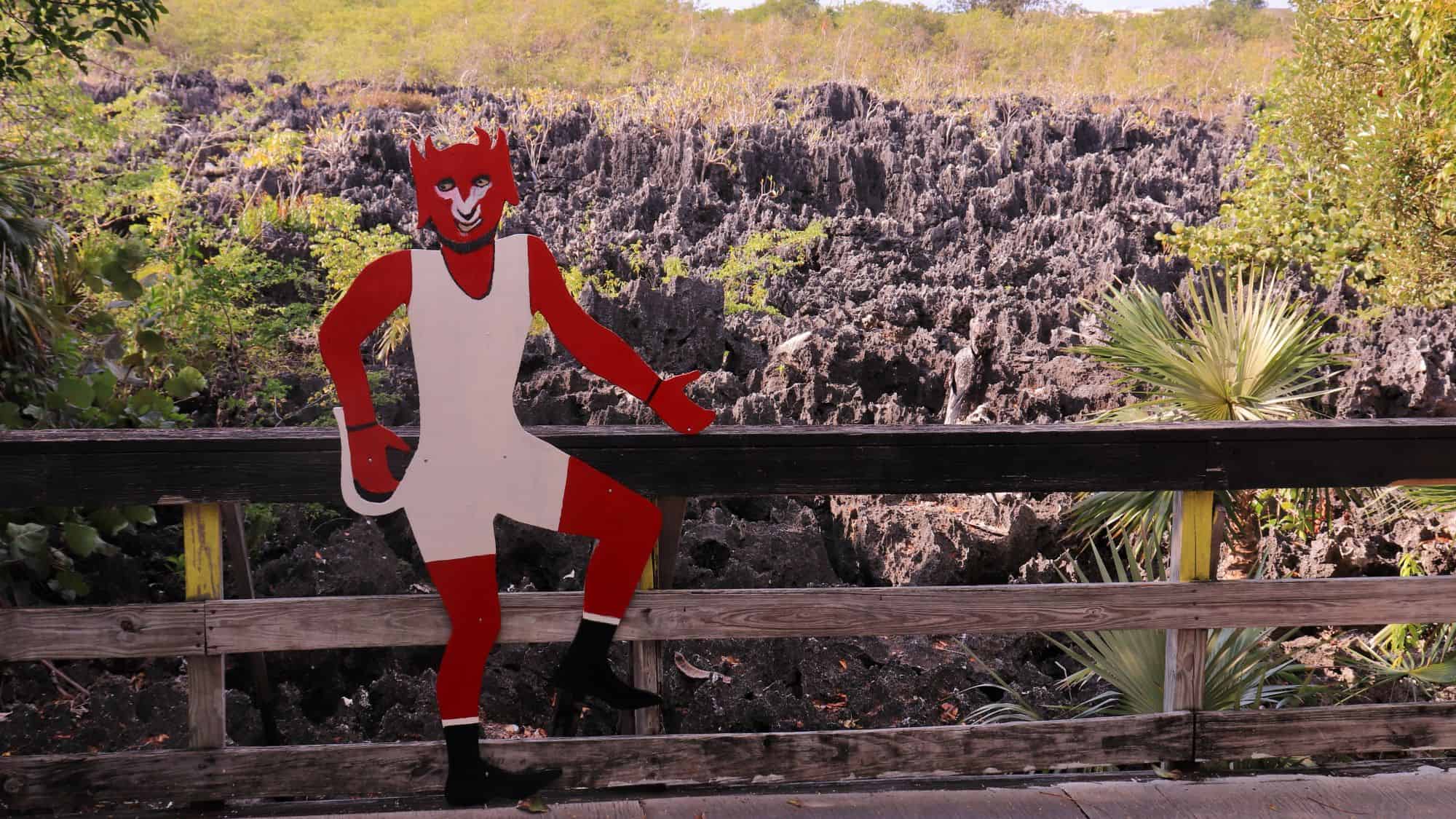 A wooden boardwalk overlooks jagged black limestone formations, resembling a scene from another world. A red devil cutout with a grin leans against the railing, marking the famous "Hell" tourist attraction in the Cayman Islands.