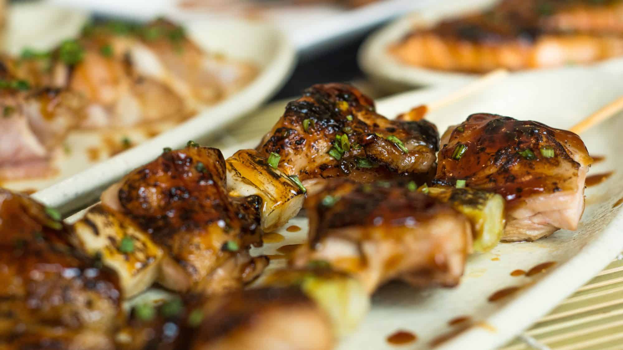 A close-up of grilled salmon skewers with a caramelized glaze, served on a plate with green herbs sprinkled on top. The skewers feature alternating pieces of salmon and grilled vegetables, with more salmon dishes visible in the blurred background.